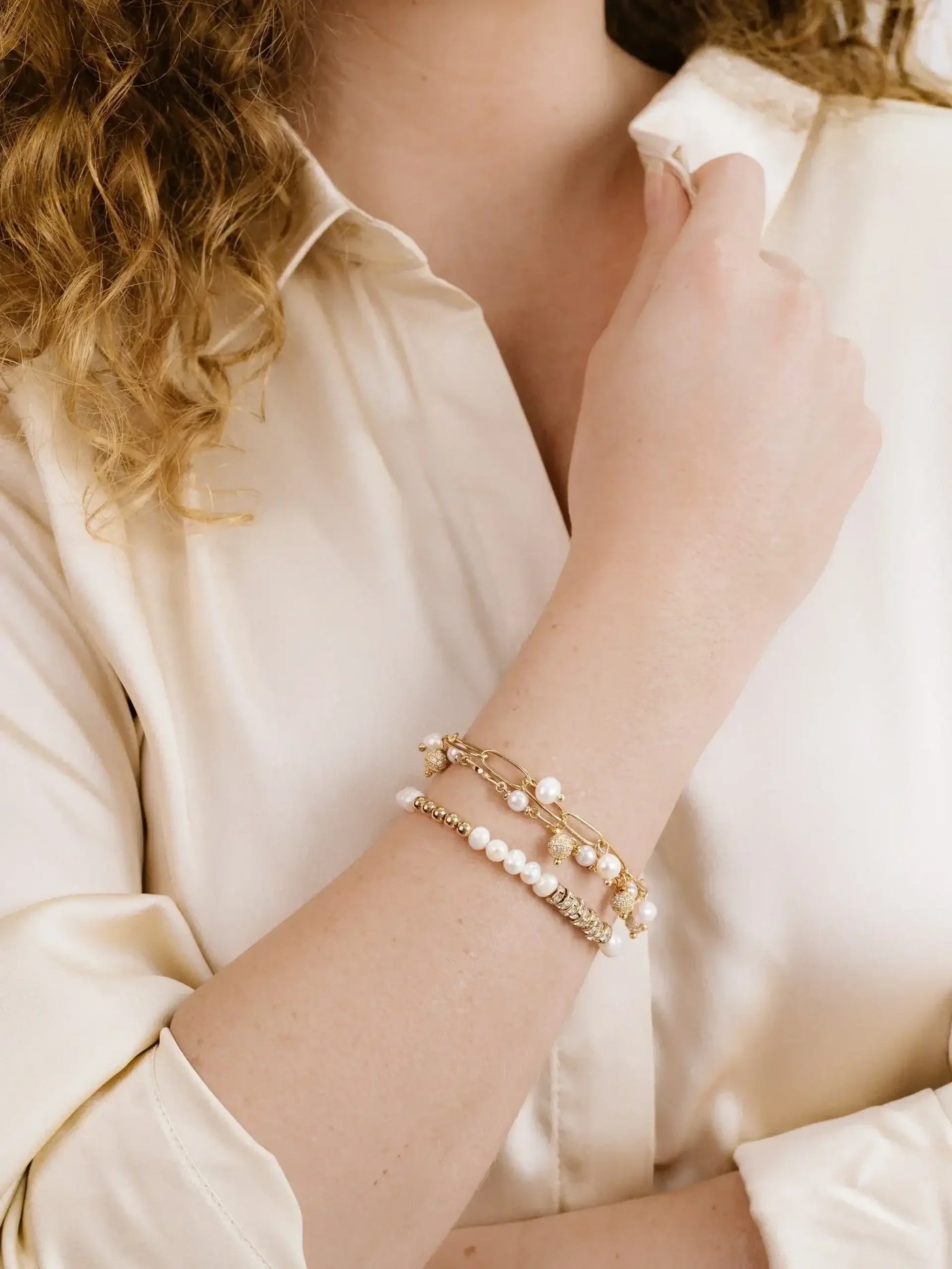A hand with multiple elegant bracelets rests against a light, draped fabric. The bracelets feature gold and pearl accents, complementing a soft cream-colored blouse.