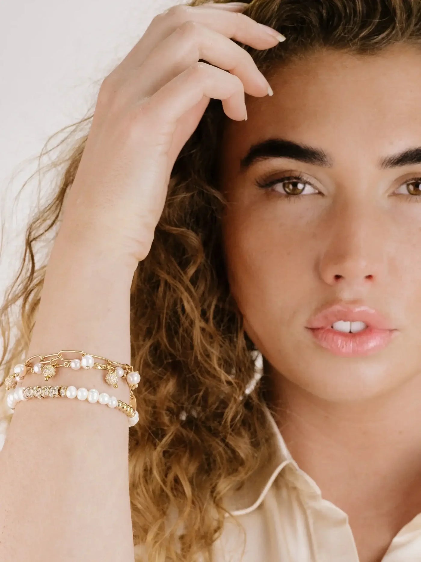 A woman with curly hair touches her hair while showcasing multiple bracelets on her wrist. She has a natural makeup look, and the background is neutral and soft.