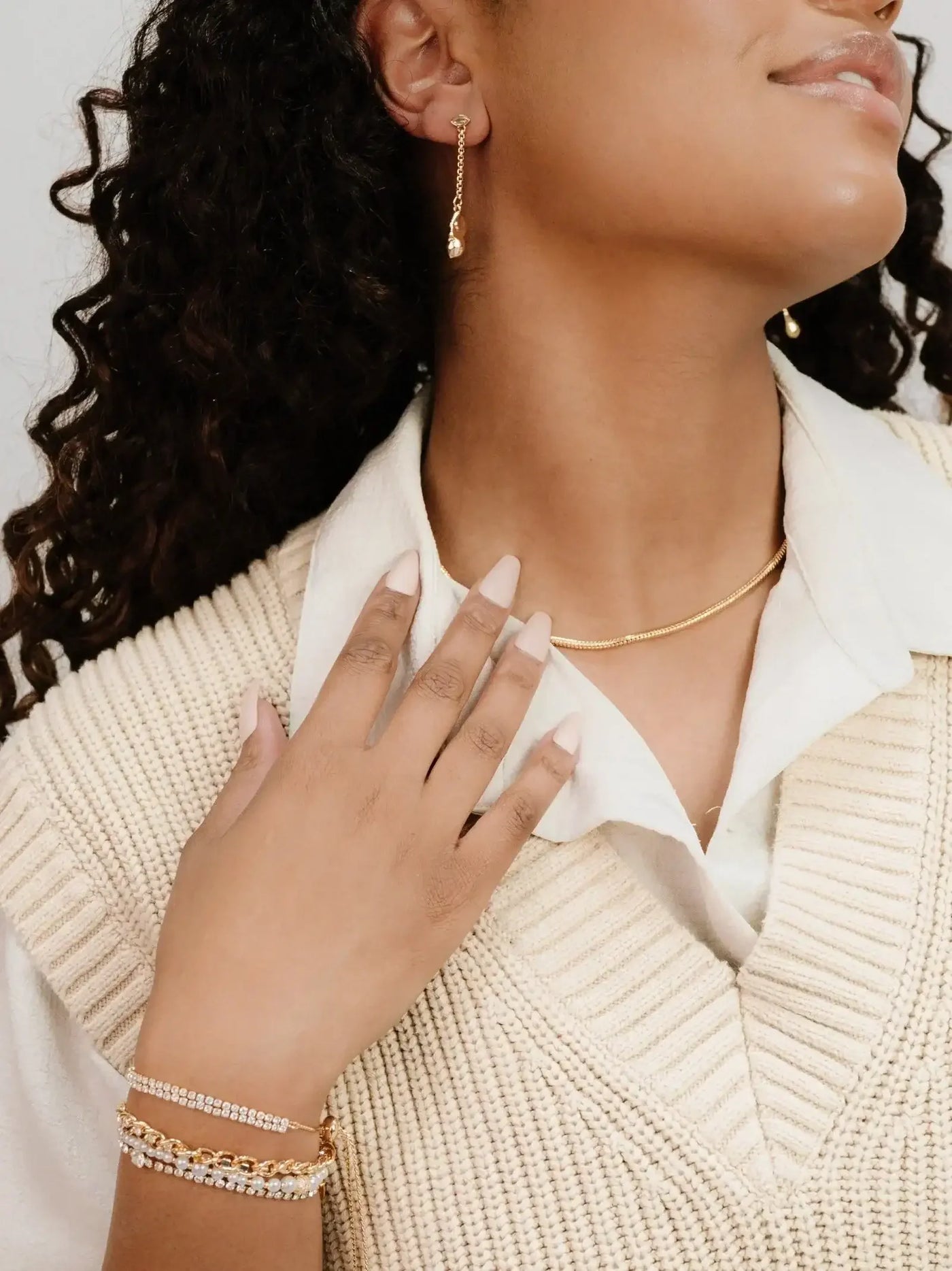 A woman with curly hair wears gold earrings and layered bracelets, gently touching her neck. She is dressed in a beige knitted vest over a white shirt, positioned against a light background.
