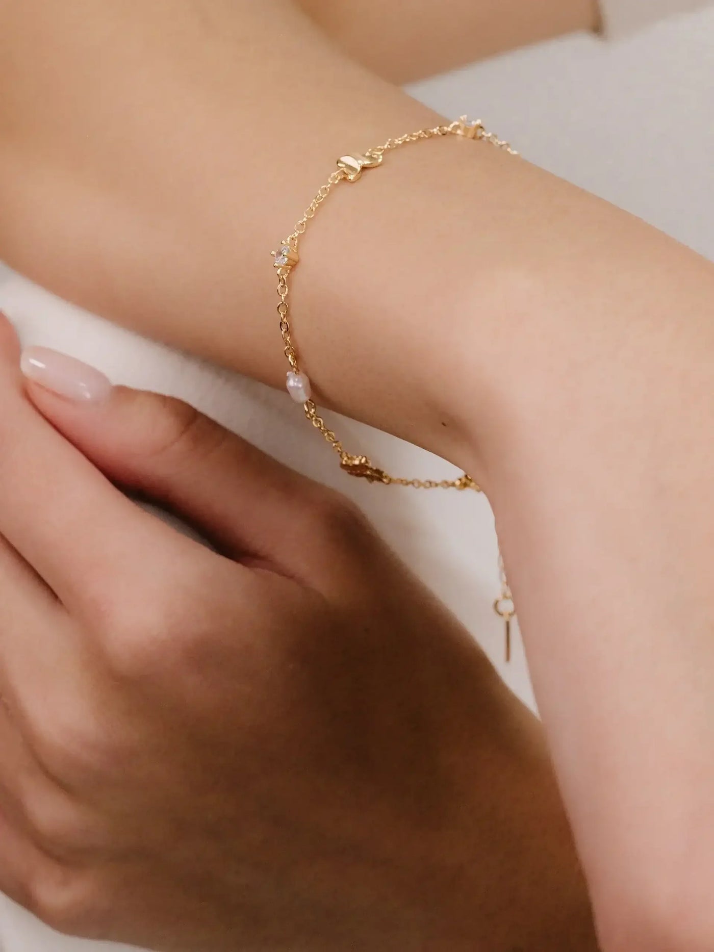 A delicate gold bracelet adorned with small pearls and shiny accents rests on a woman's wrist, while her other hand gently holds it against a soft, light-colored background.