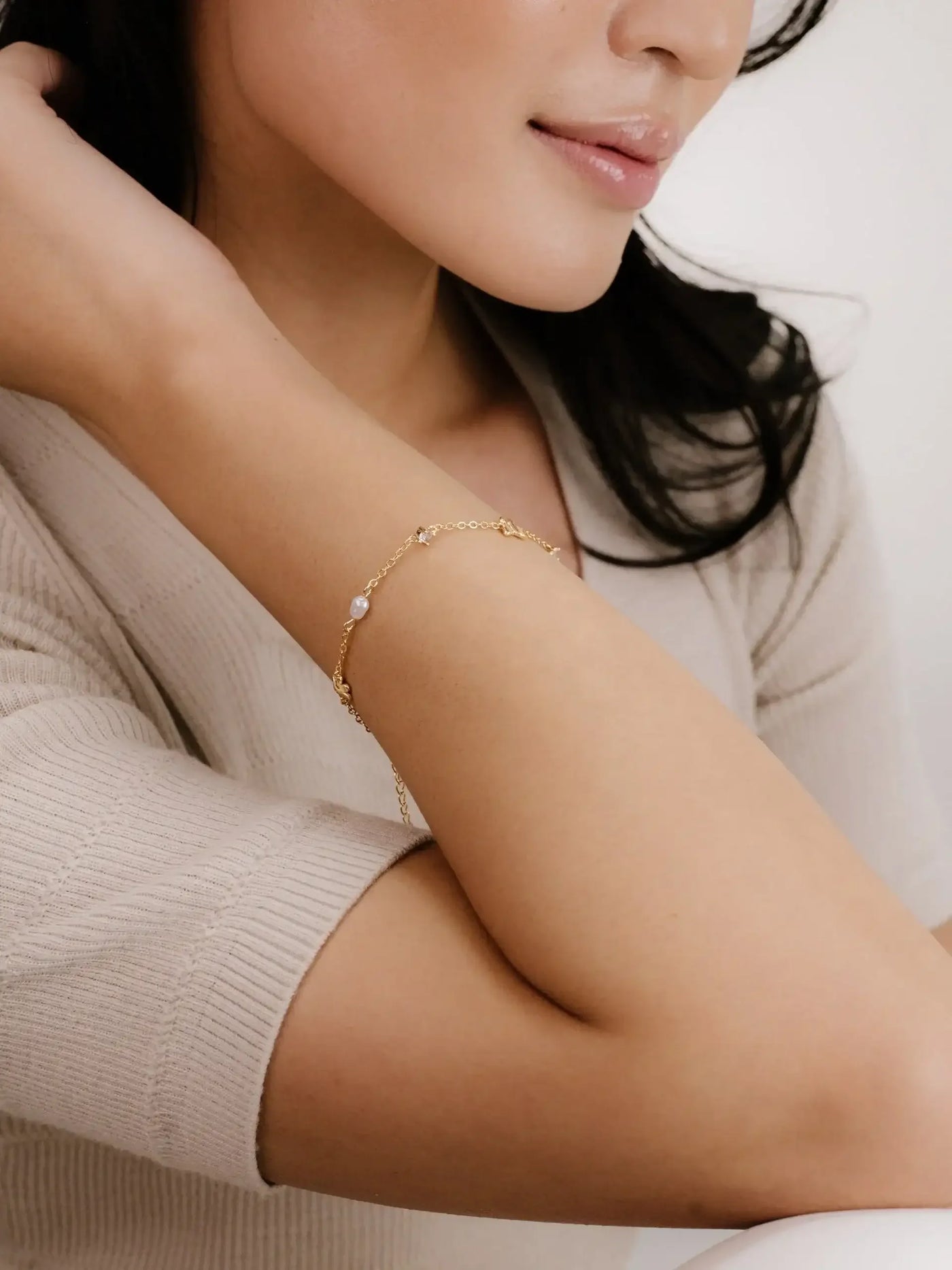 A gold bracelet with pearls is elegantly worn on a woman's arm, while she tilts her head slightly, showcasing a soft, serene expression against a light, neutral background.
