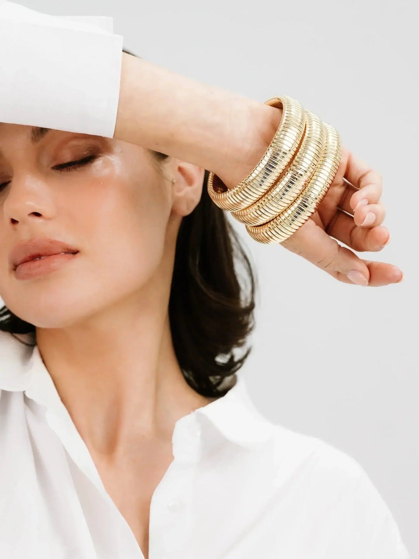 A woman with a relaxed expression wears multiple shiny golden bangles on her arm while touching her forehead. She is dressed in a white shirt against a neutral background.