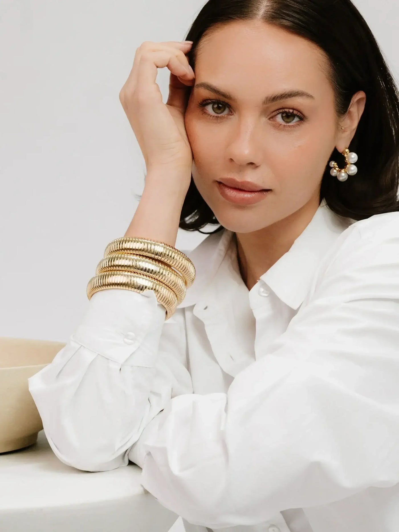 A woman in a white shirt rests her chin on her hand, showcasing multiple gold bangles and pearl earrings. She is seated beside a circular, light-colored table in a minimalistic setting.