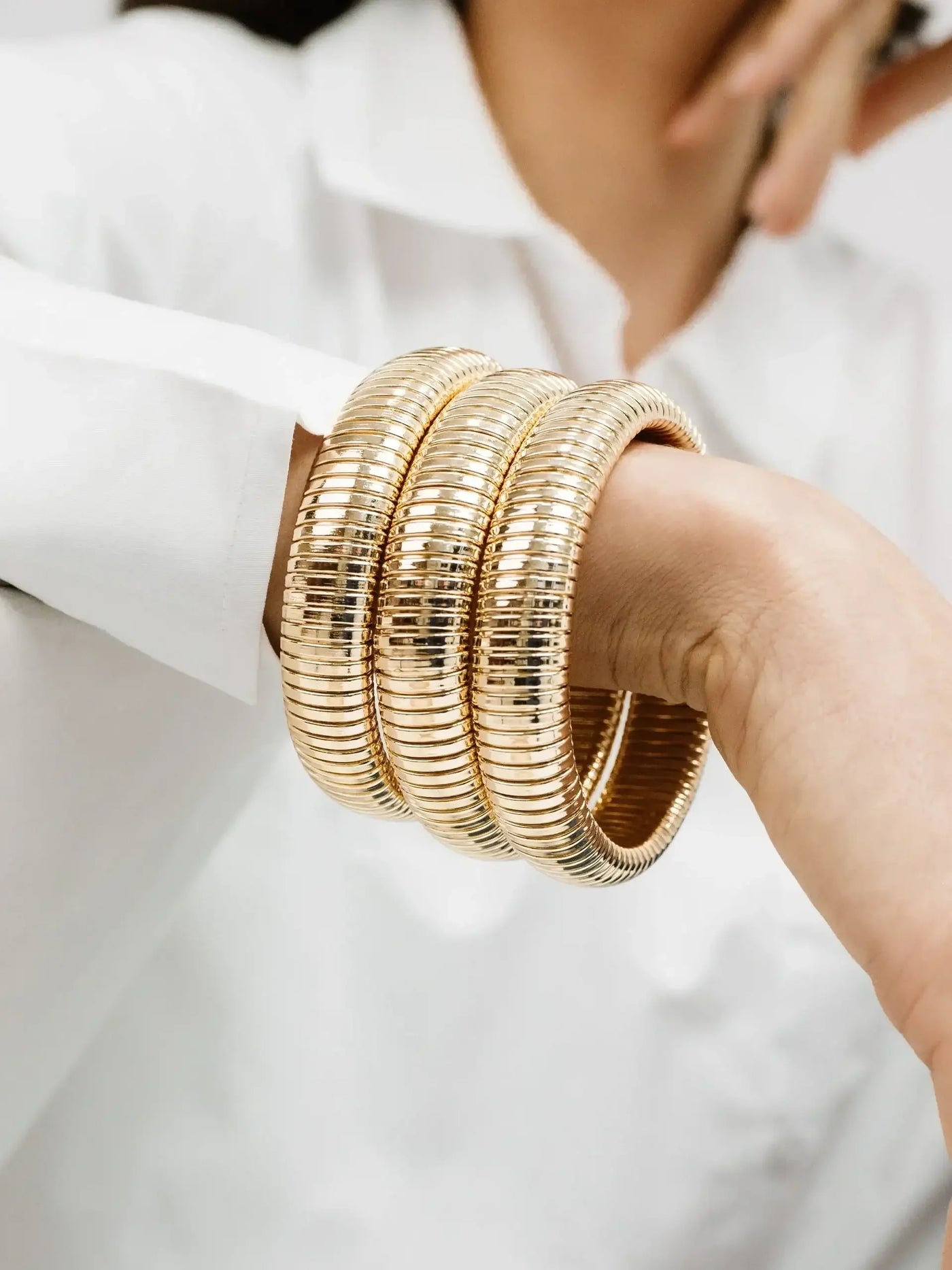Three shiny gold bangles are stacked on a wrist. The hand is slightly raised, showcasing the jewelry. The background features a light-colored, blurred setting, emphasizing the wrist adornments.