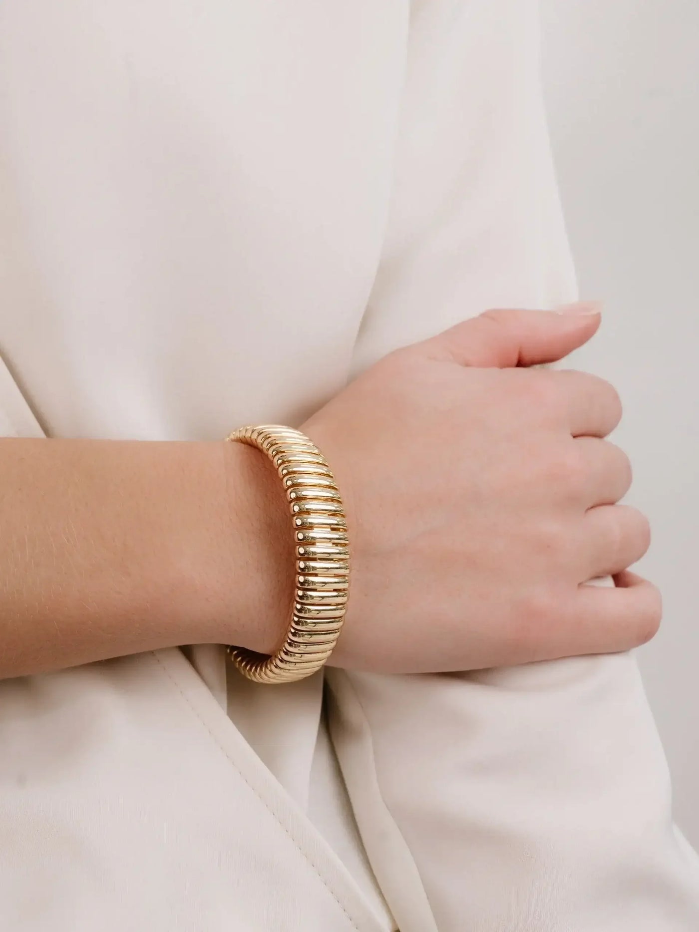 A gold bracelet adorns a person's wrist, resting on a light-colored, smooth fabric. The individualâ€™s hand is partially visible, gently grasping their arm, conveying a relaxed pose.