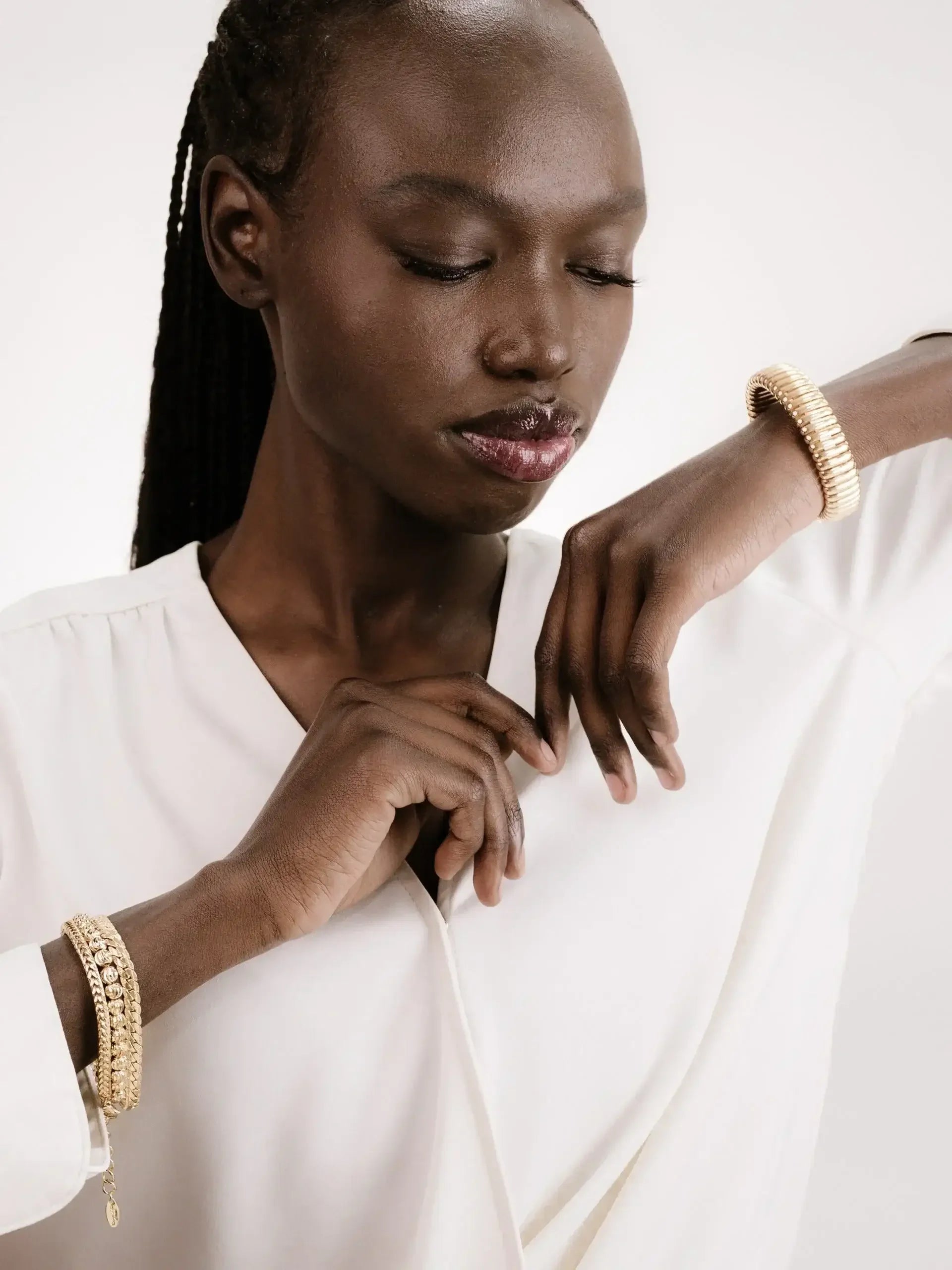 A smooth-skinned individual styled in a white blouse delicately touches their wrist adorned with multiple gold bracelets, exuding elegance against a muted, neutral backdrop.