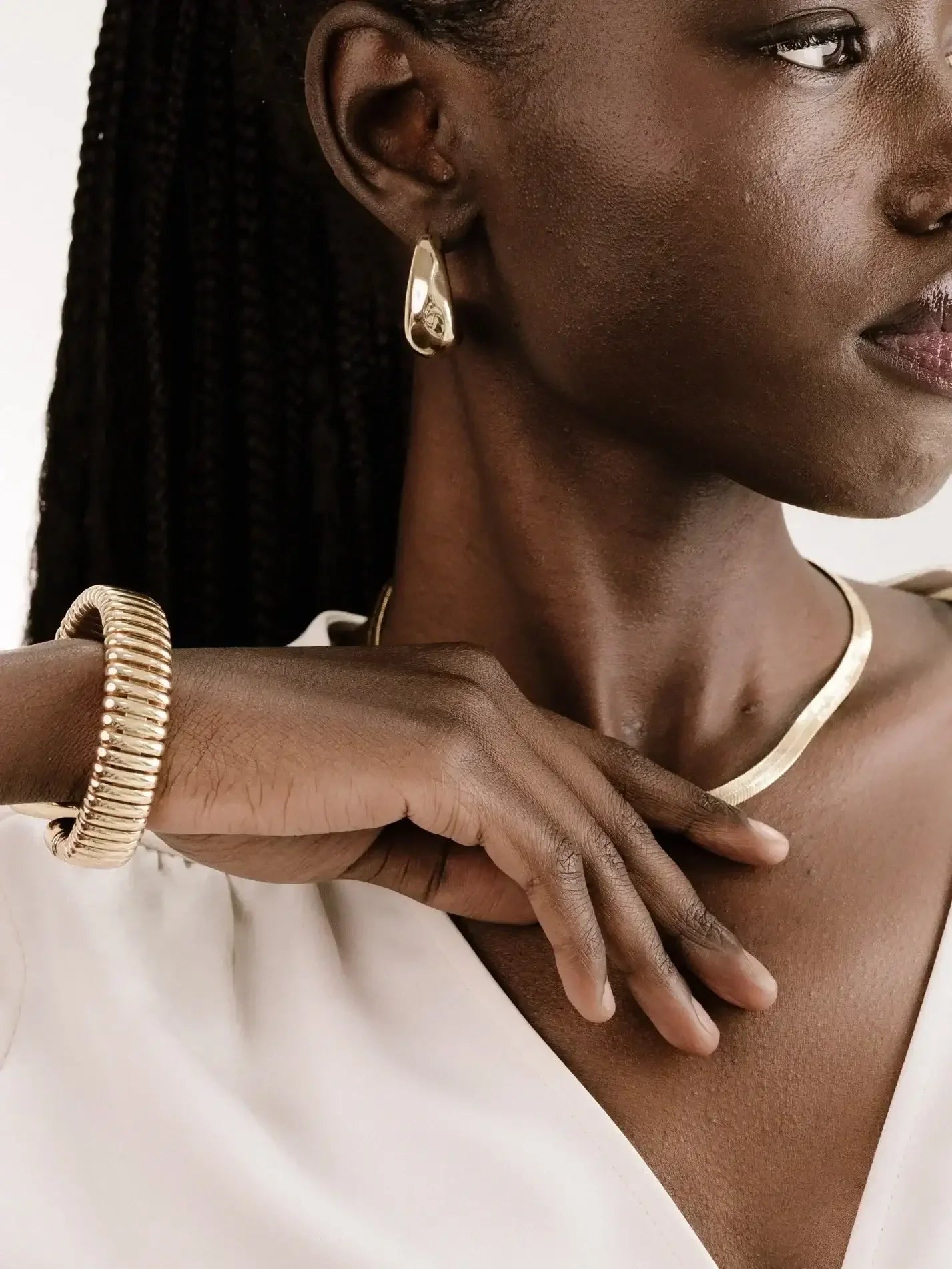 A person with dark skin and braided hair is elegantly showcasing gold jewelry, including earrings and a bracelet, while gently placing their hand on their collarbone, against a neutral background.