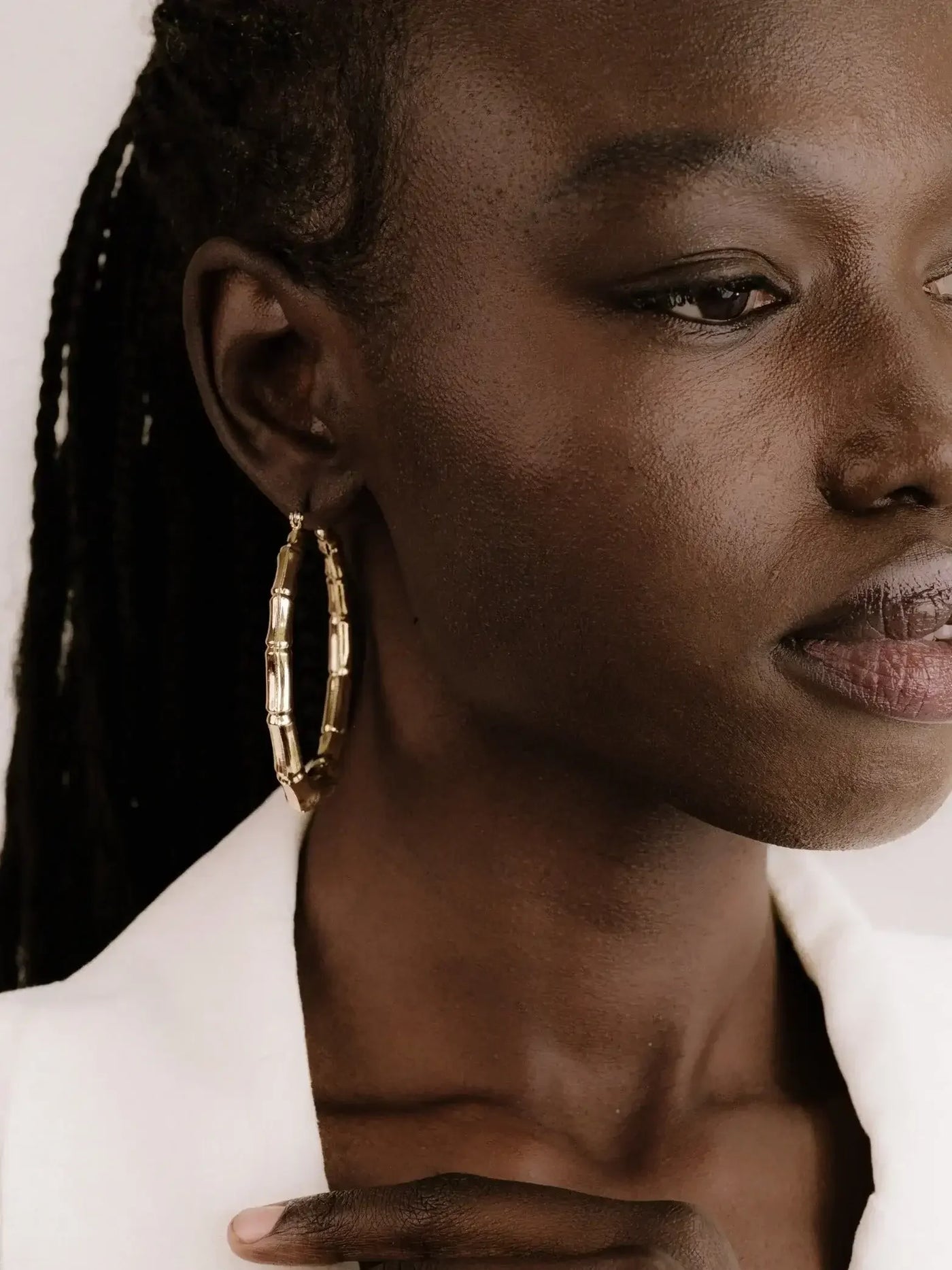 A close-up of a dark-skinned woman wearing large, golden hoop earrings, gently touching her collarbone with one hand while looking thoughtfully to the side against a soft, neutral background.