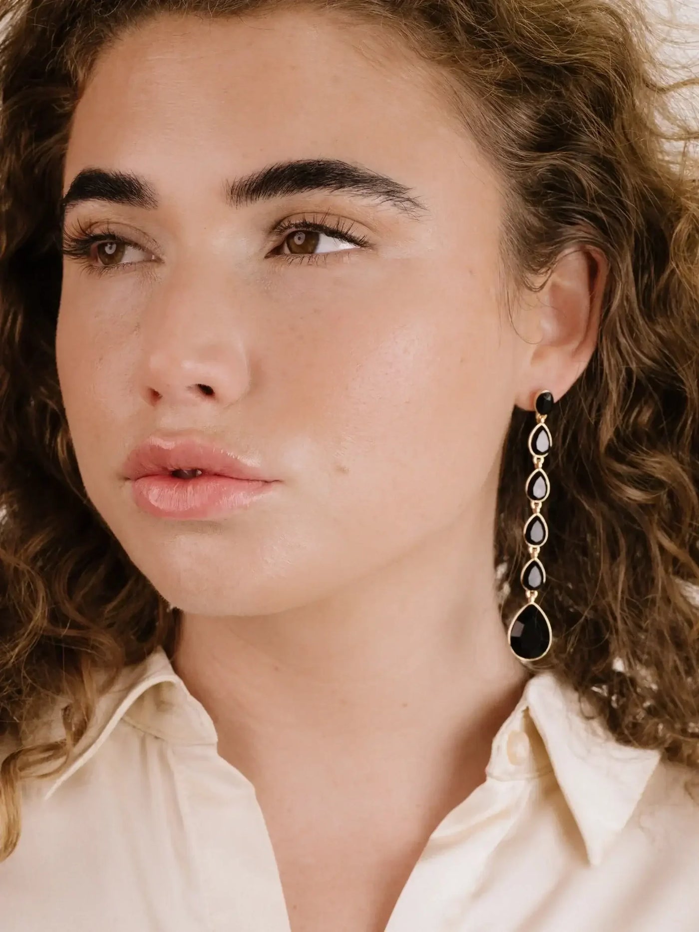 A woman with curly hair and a pale complexion is wearing a long, decorative earring. She gazes to the side, dressed in a light-colored, button-up shirt against a neutral background.
