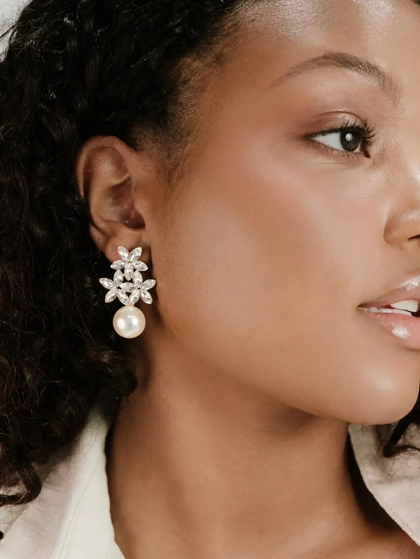 A woman with curly hair wears ornate earrings featuring flowers and a pearl. She gazes slightly to the side, showcasing her elegant accessories against a muted background.