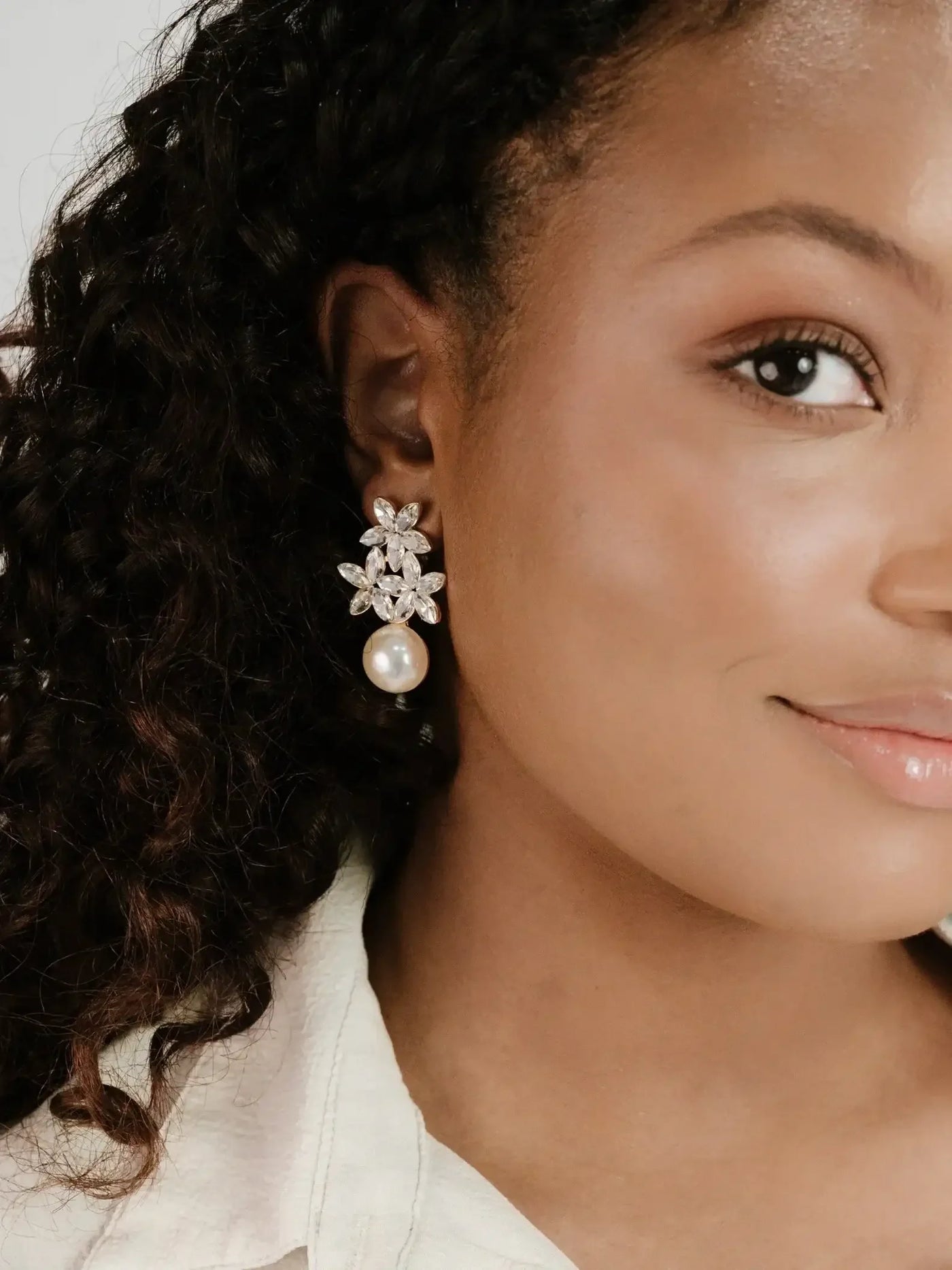 A close-up of a woman's face showcases her wearing an ornate earring featuring a floral design with sparkling stones and a pearl drop, set against a neutral background.