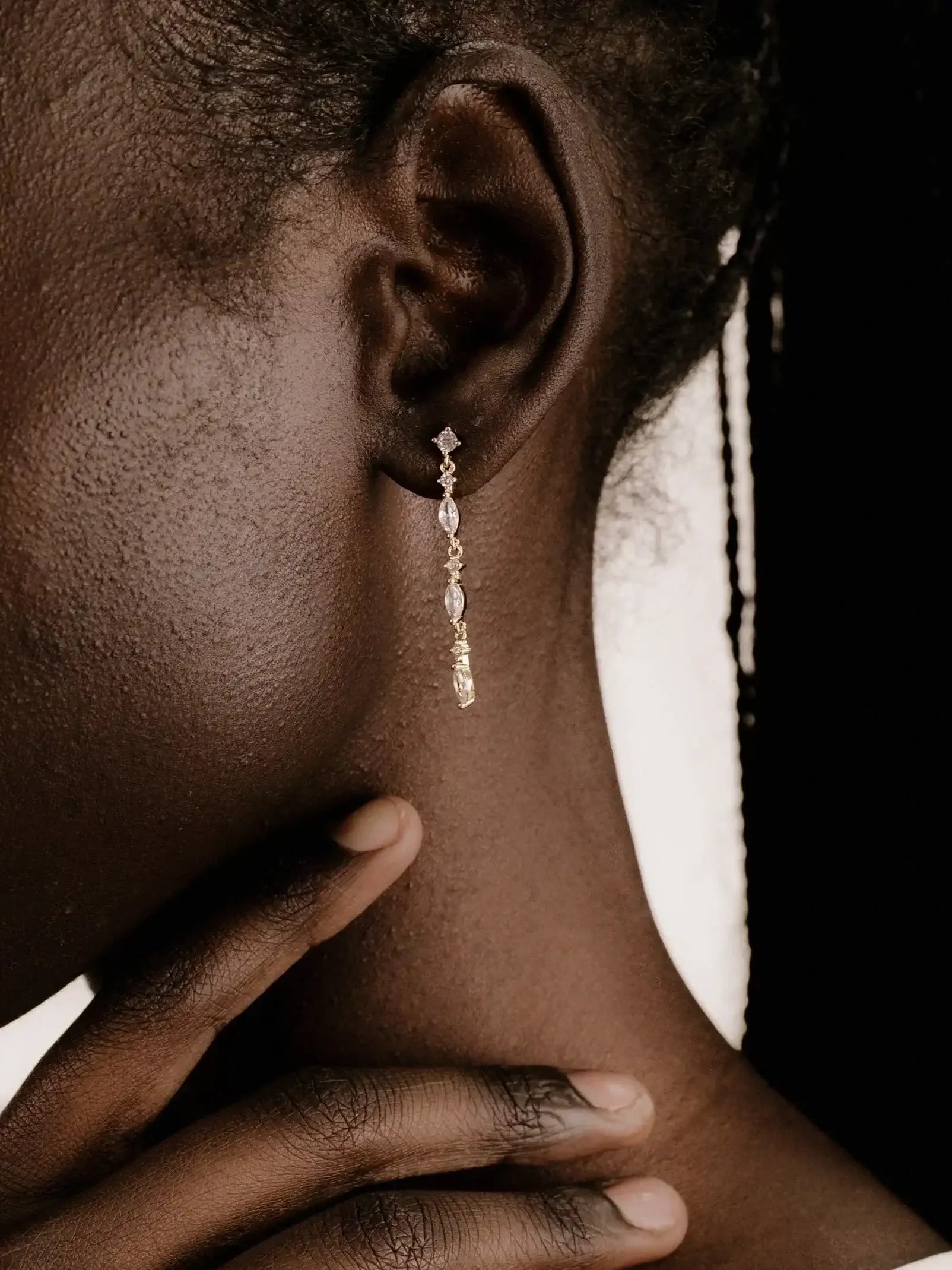 A close-up of a person's ear adorned with a dangling gold earring features a gemstone. The individual gently touches their neck with one hand, emphasizing the elegant jewelry against dark skin and a neutral background.