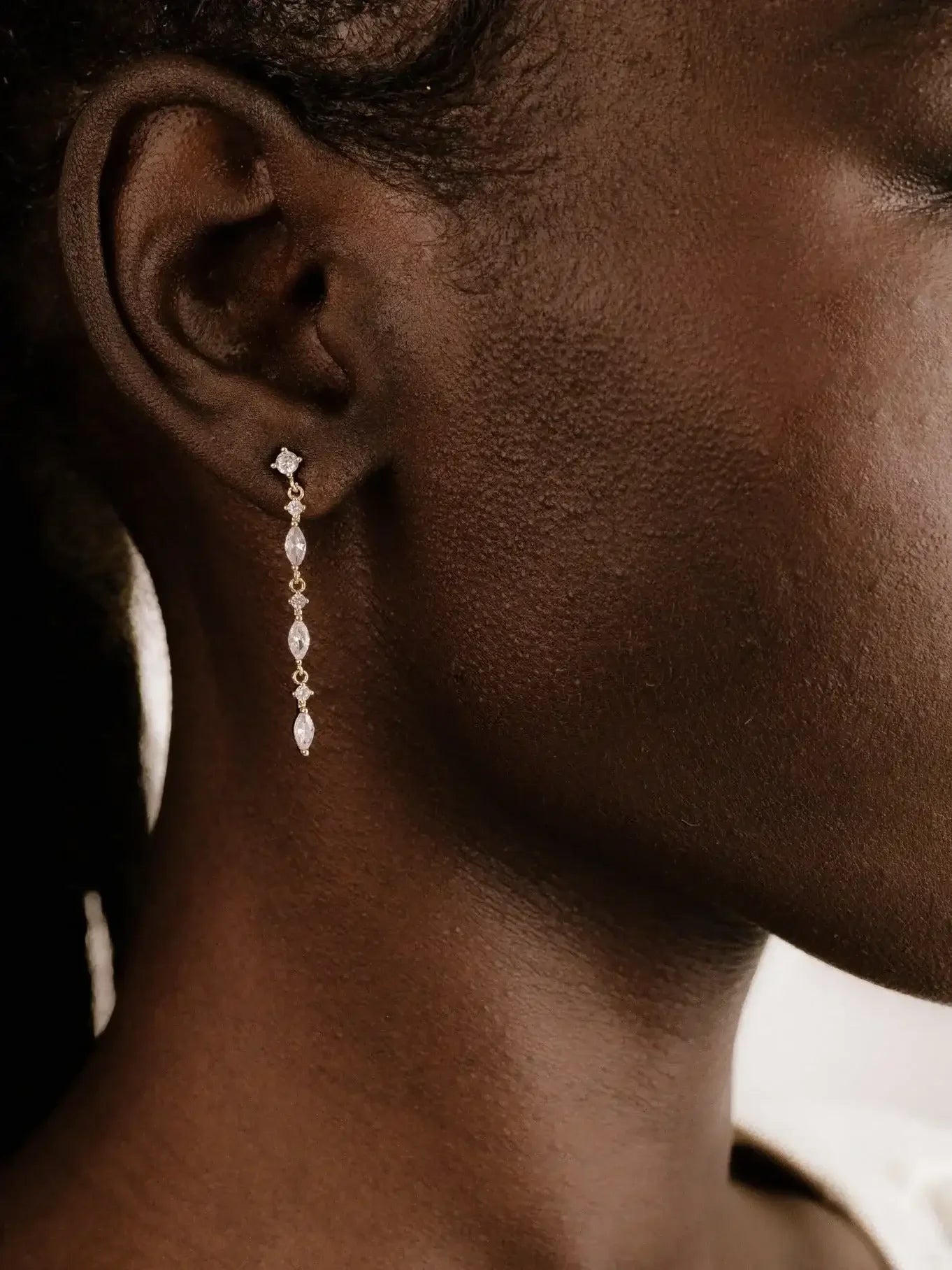 A delicate earring adorns the lobe of a dark-skinned person's ear, glinting with small gemstones, against a softly blurred background that suggests a neutral and minimalist setting.