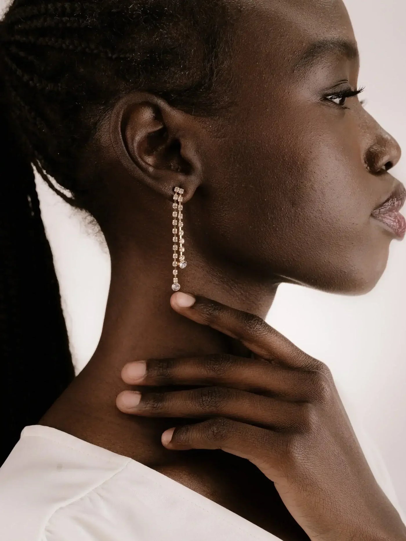 A profile of a woman with braided hair touches her neck, adorned with a dangling earring featuring small, shiny beads. The background is neutral and softly lit.