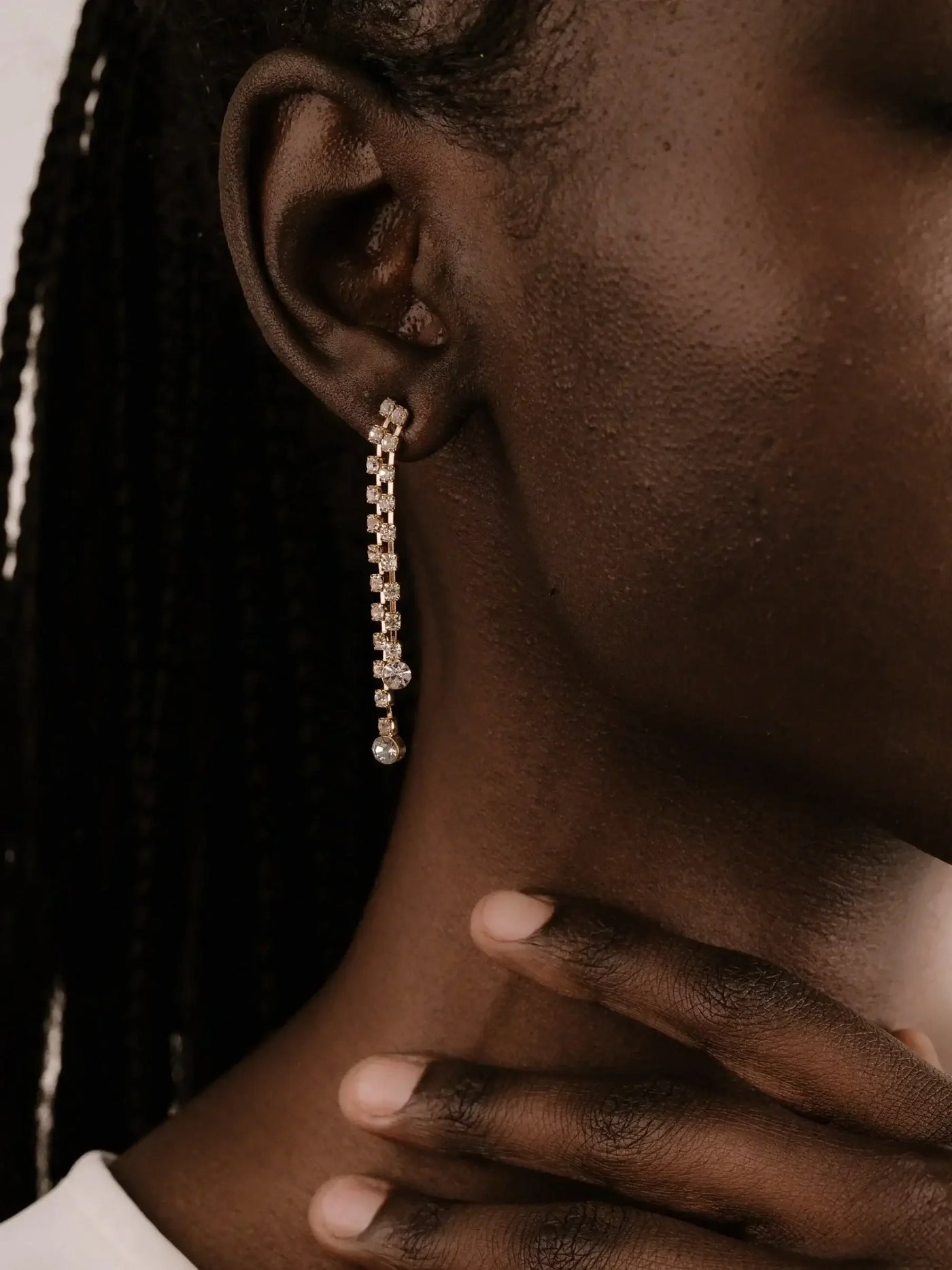A gold dangle earring with sparkly elements is worn on an ear. The hand gently touches the neck, highlighting the earring, against a neutral background.