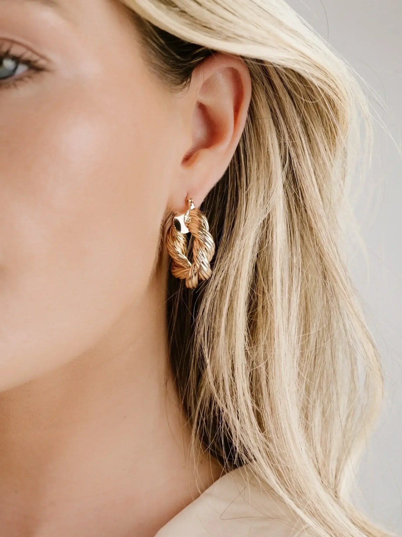 A gold hoop earring in a twisted design hangs from a modelâ€™s ear, accentuating her light blonde hair and soft skin in a neutral-toned setting.