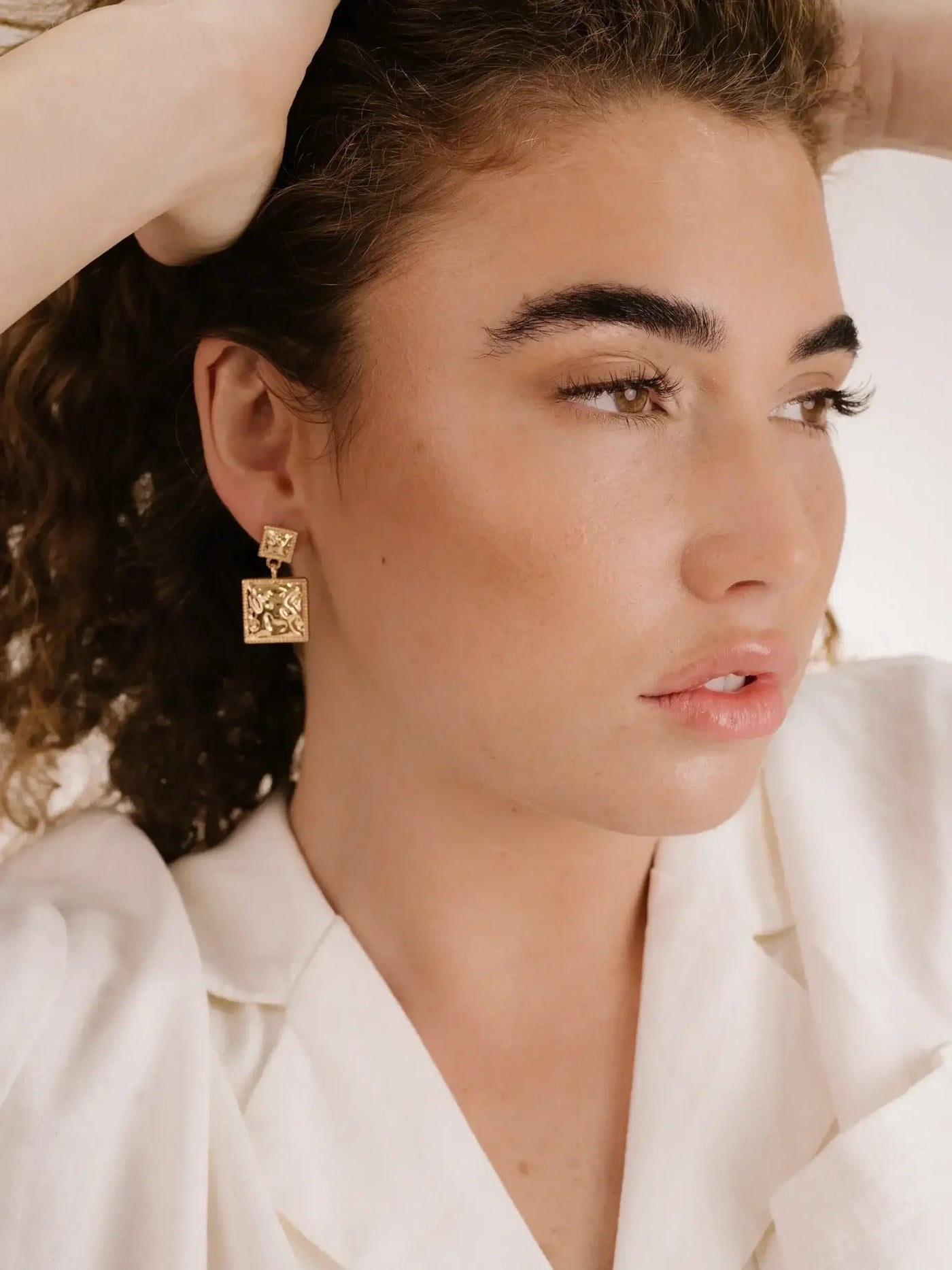 A young woman with curly hair poses thoughtfully, wearing large gold earrings. She gently holds her hair back with one hand, showcasing her neutral-toned outfit against a soft, light background.