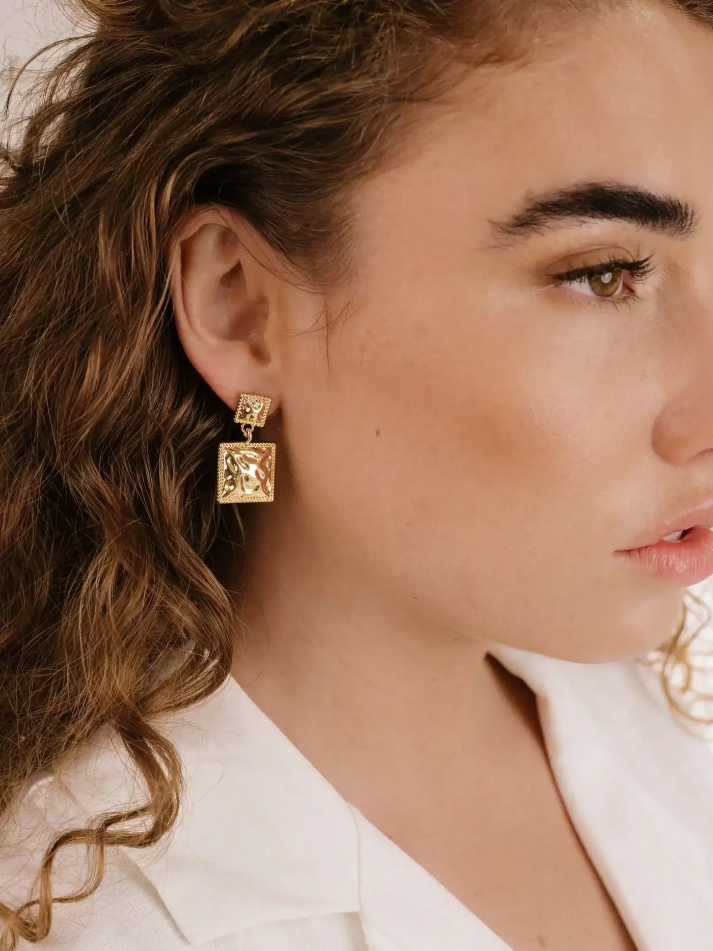 A woman with curly hair wears stylish gold earrings featuring square designs. The earrings hang closely to her face in a soft, neutral-toned setting, highlighting her elegant features.