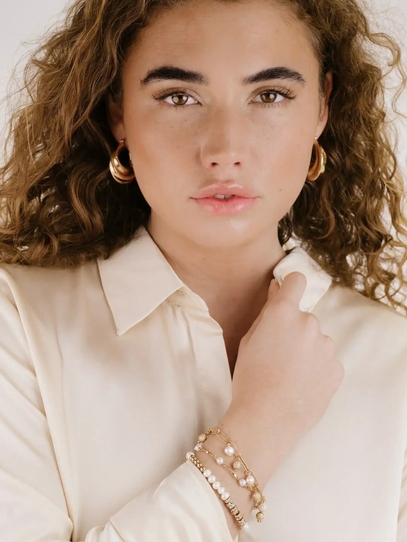 A young woman with curly hair poses, wearing a cream-colored blouse and gold hoop earrings. She gently touches her neck with one hand, showcasing multiple bracelets on her wrist.