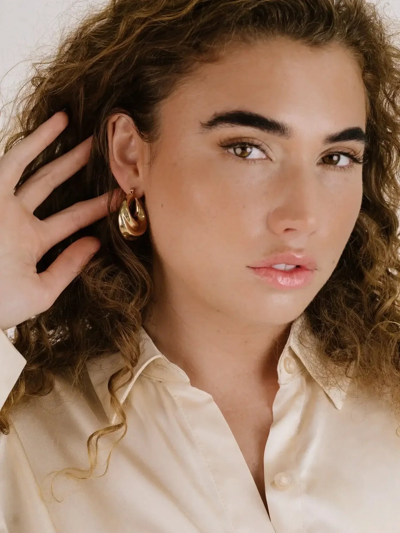 A young woman with curly hair holds her ear, showcasing a large, shiny gold earring. She wears a light, collared, satin shirt and gazes directly at the camera, exuding a confident expression.