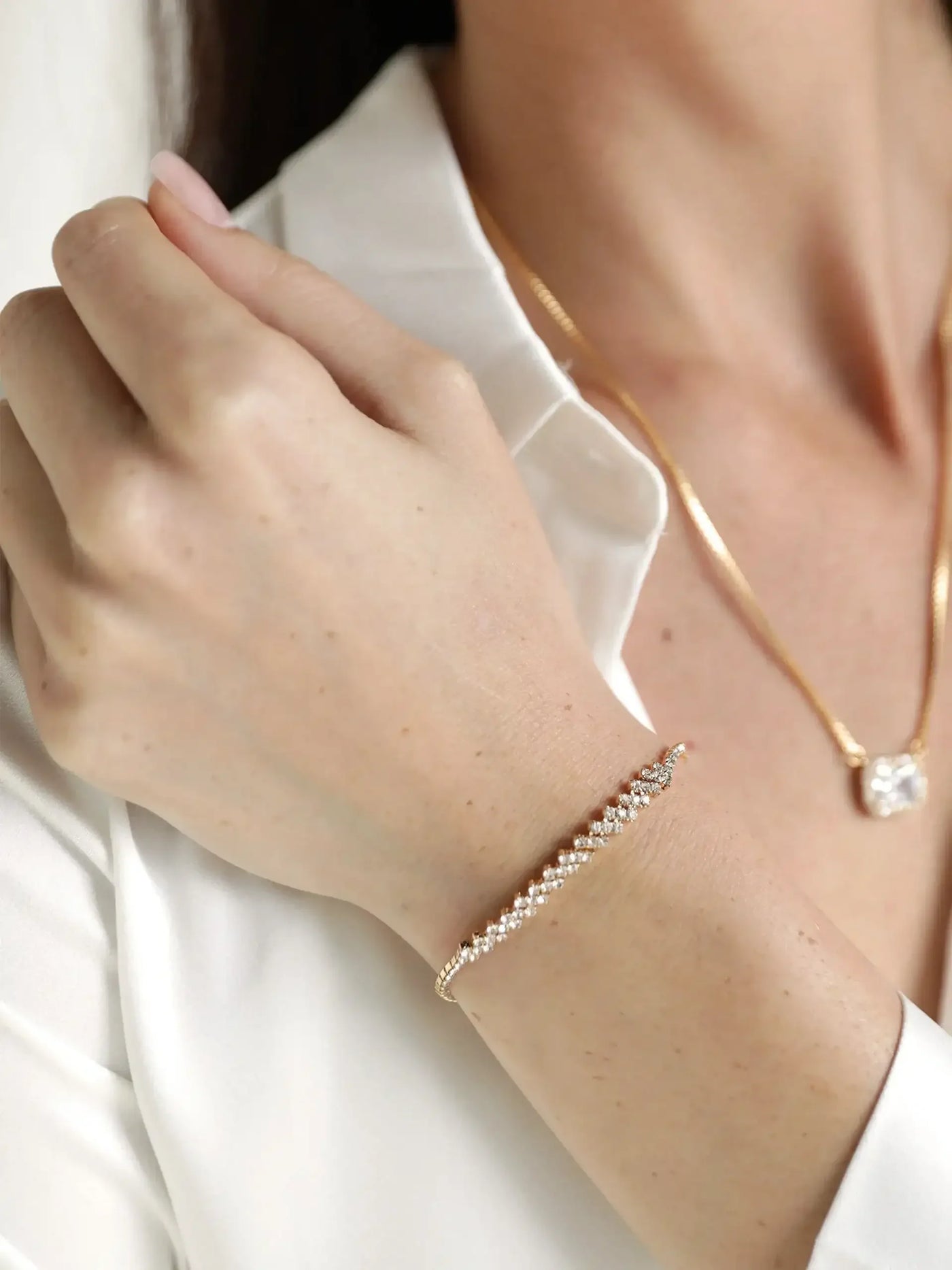 A hand adorned with a delicate, sparkling bracelet gently rests against a white blouse, complementing a gold necklace. The setting appears to be neutral and elegant.