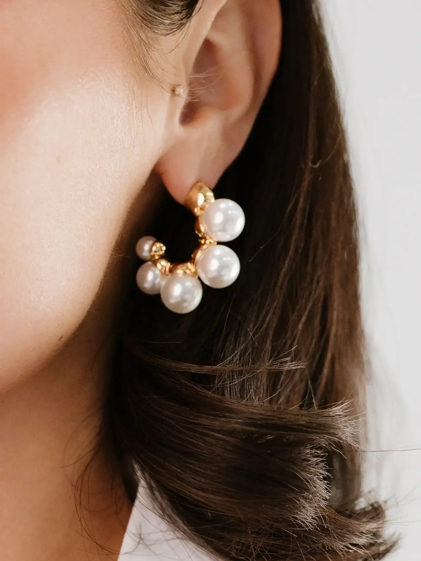 A close-up of a woman's ear showcasing a chunky gold earring adorned with multiple large white pearls, set against a soft, neutral background, emphasizing the elegant accessory.