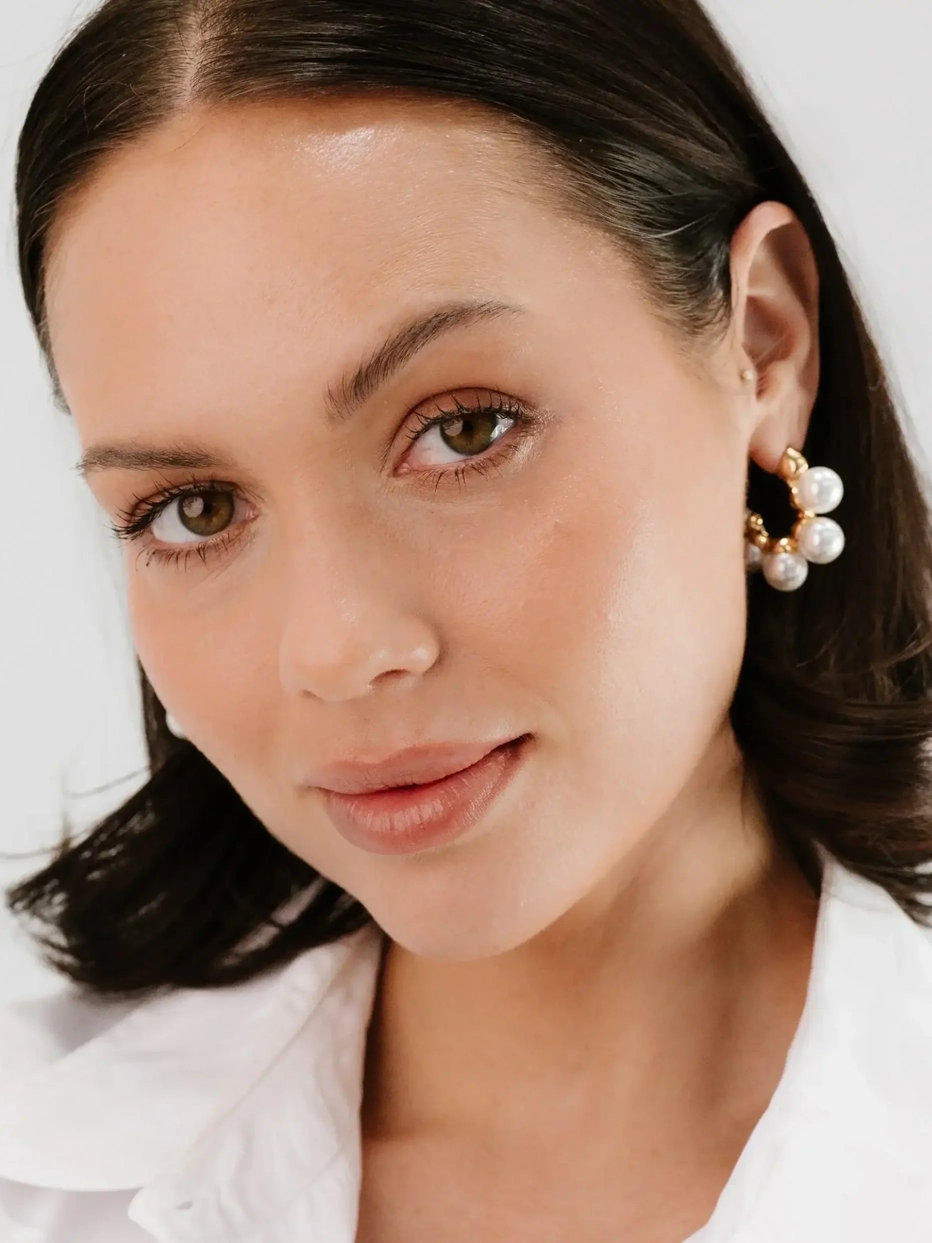 A woman with brown eyes and natural makeup is smiling, wearing fashionable pearl earrings, set against a clean, bright background while partially tucked within a white collared shirt.