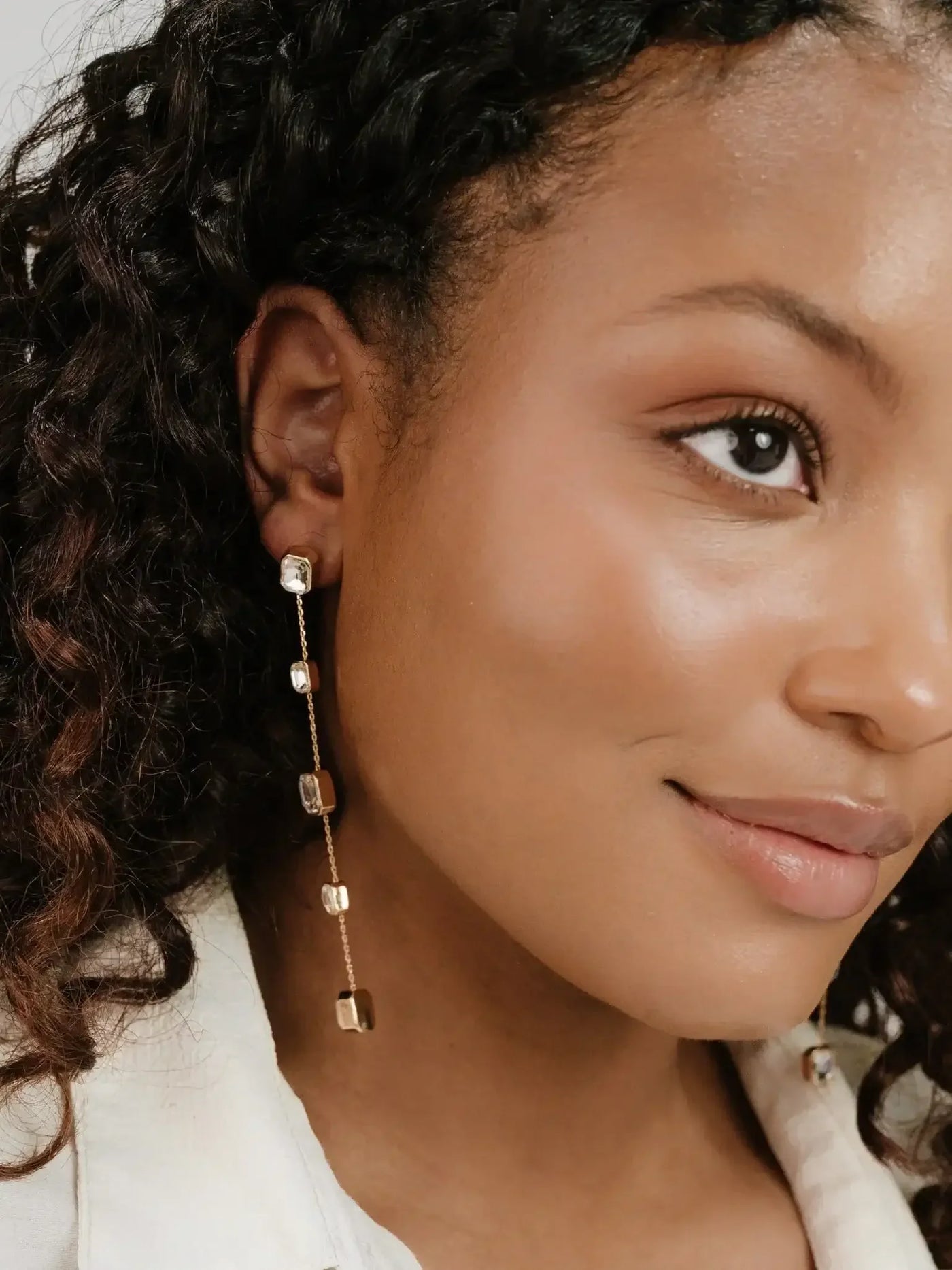 A woman with curly hair wears elegant, dangling earrings featuring gold and clear crystals. She smiles gently against a neutral background, highlighting her natural beauty and the jewelry's sparkle.