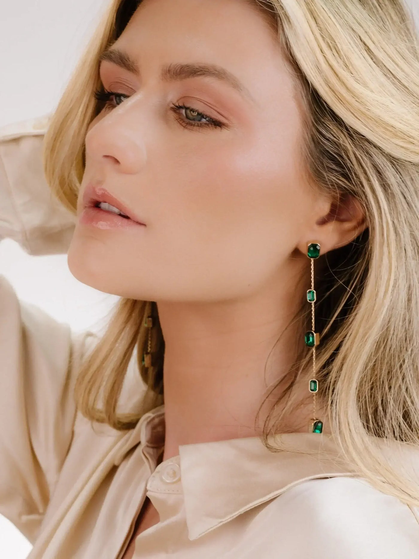 A woman with long, wavy blonde hair is delicately posed, showcasing elegant dangling emerald earrings. She wears a soft, cream-colored blouse in a simple, neutral background.