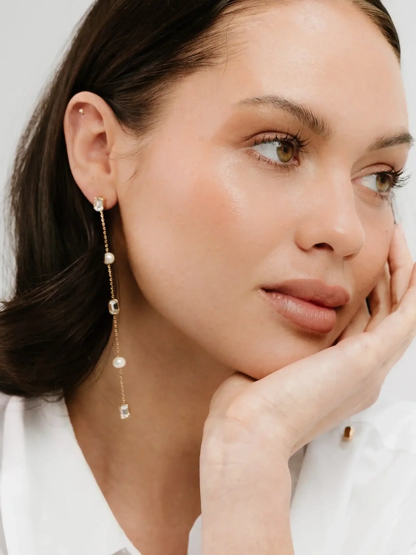 A woman with a natural makeup look rests her chin on her hand, wearing elegant, dangling earrings with pearls and gemstones against a soft, light-colored background.