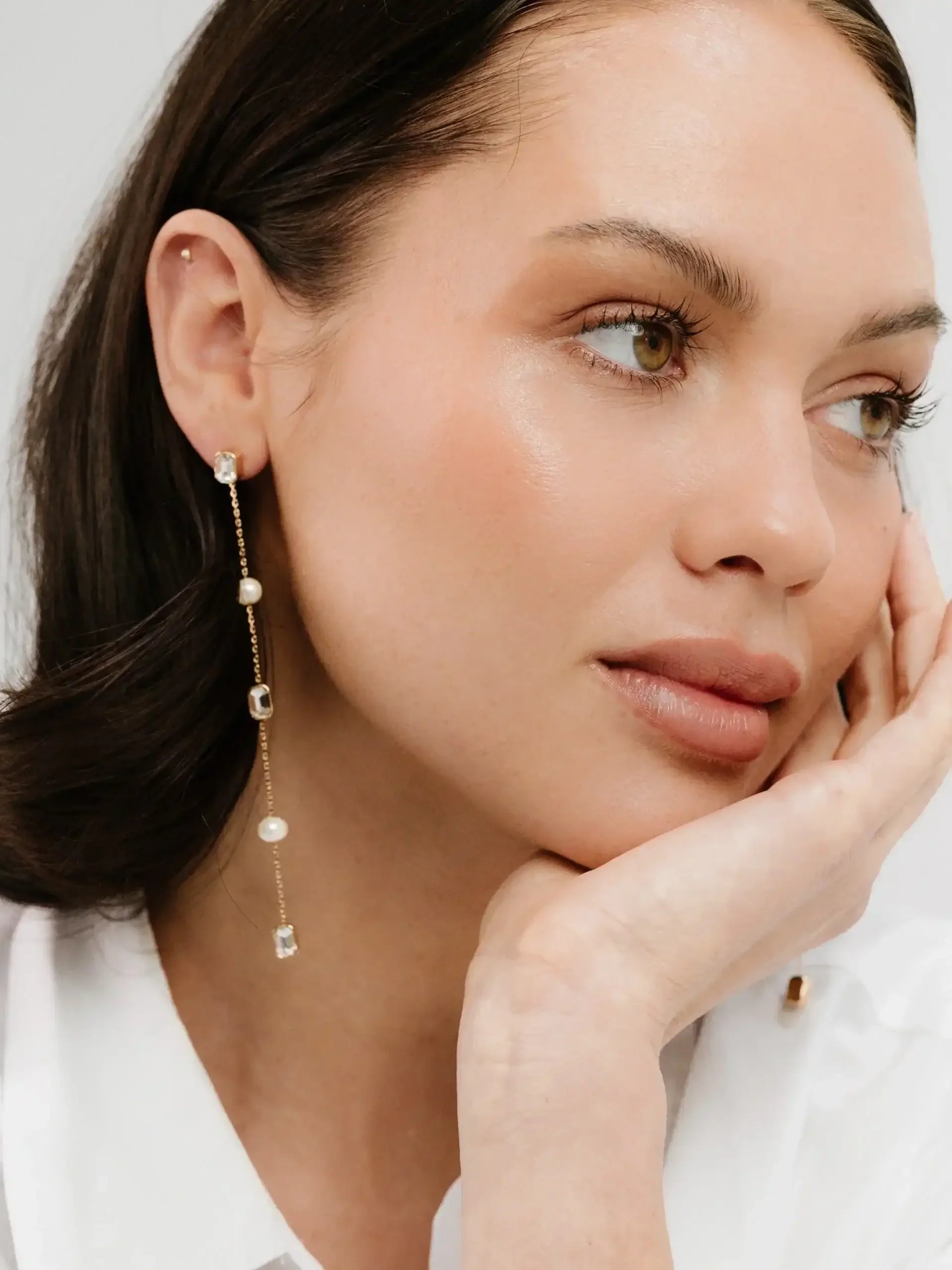 A woman with a natural makeup look rests her chin on her hand, wearing elegant, dangling earrings with pearls and gemstones against a soft, light-colored background.
