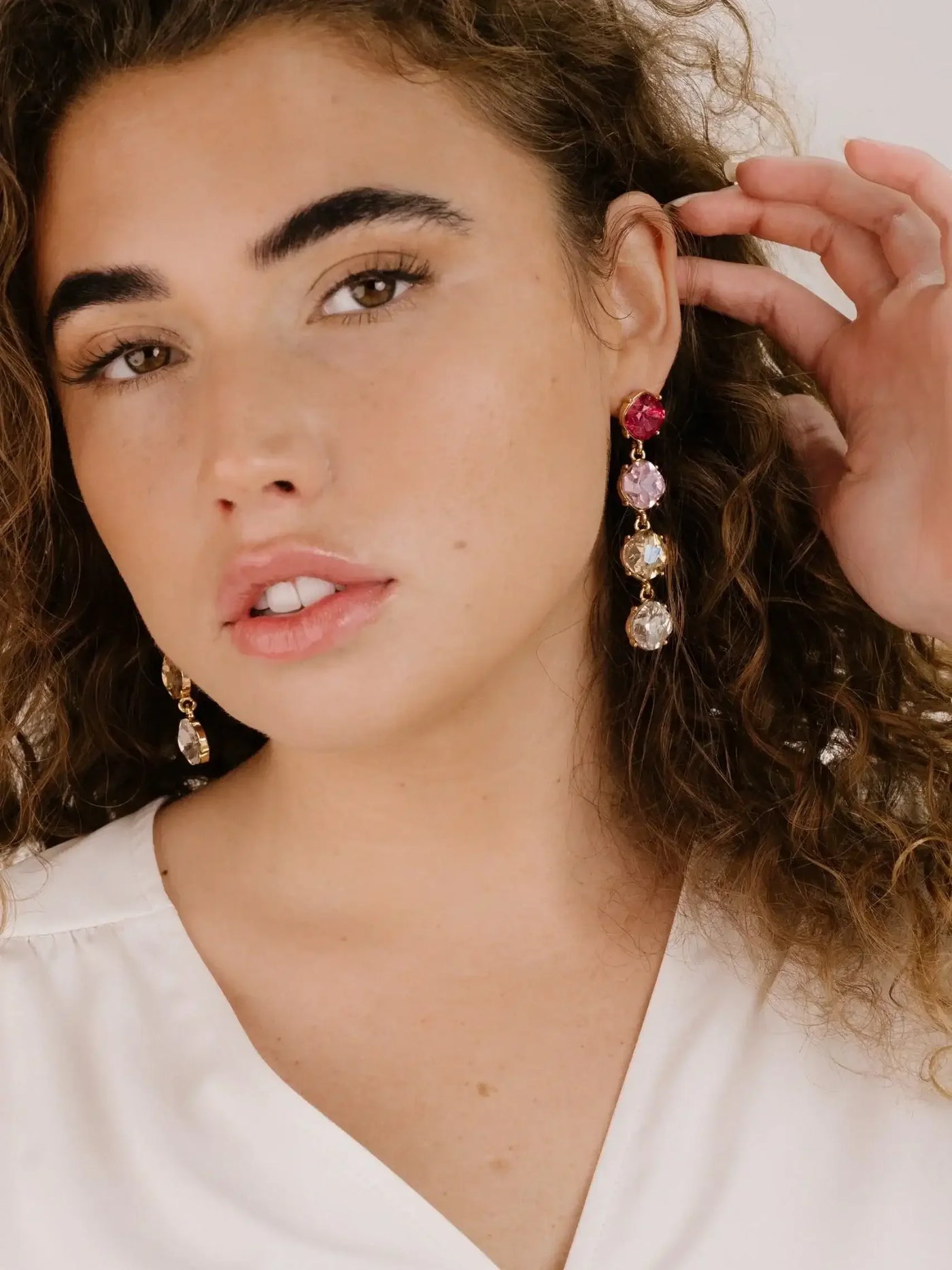 A young woman with curly hair gently touches a colorful, long earring hanging from her right ear. She wears a light blouse and has a neutral background.
