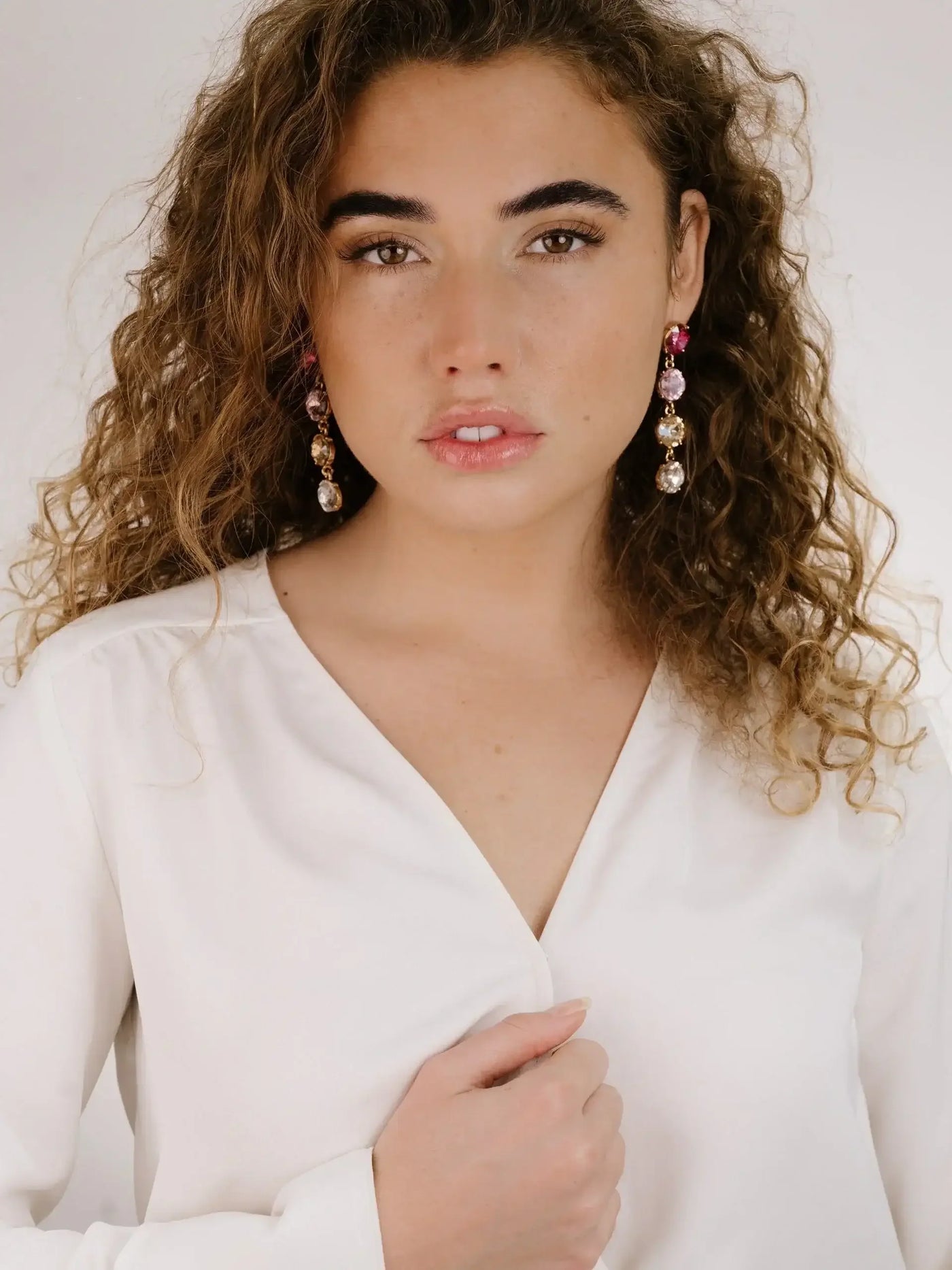 A woman with curly hair wears a white blouse and sparkling earrings. She gazes directly at the camera, with one hand gently touching her shirt, in a minimalistic studio setting.