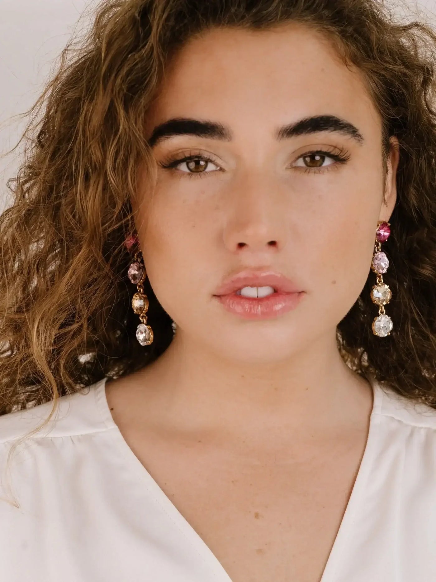A young woman with curly hair wears decorative earrings featuring various gems. She is facing the camera, displaying a natural makeup look against a simple, light background.