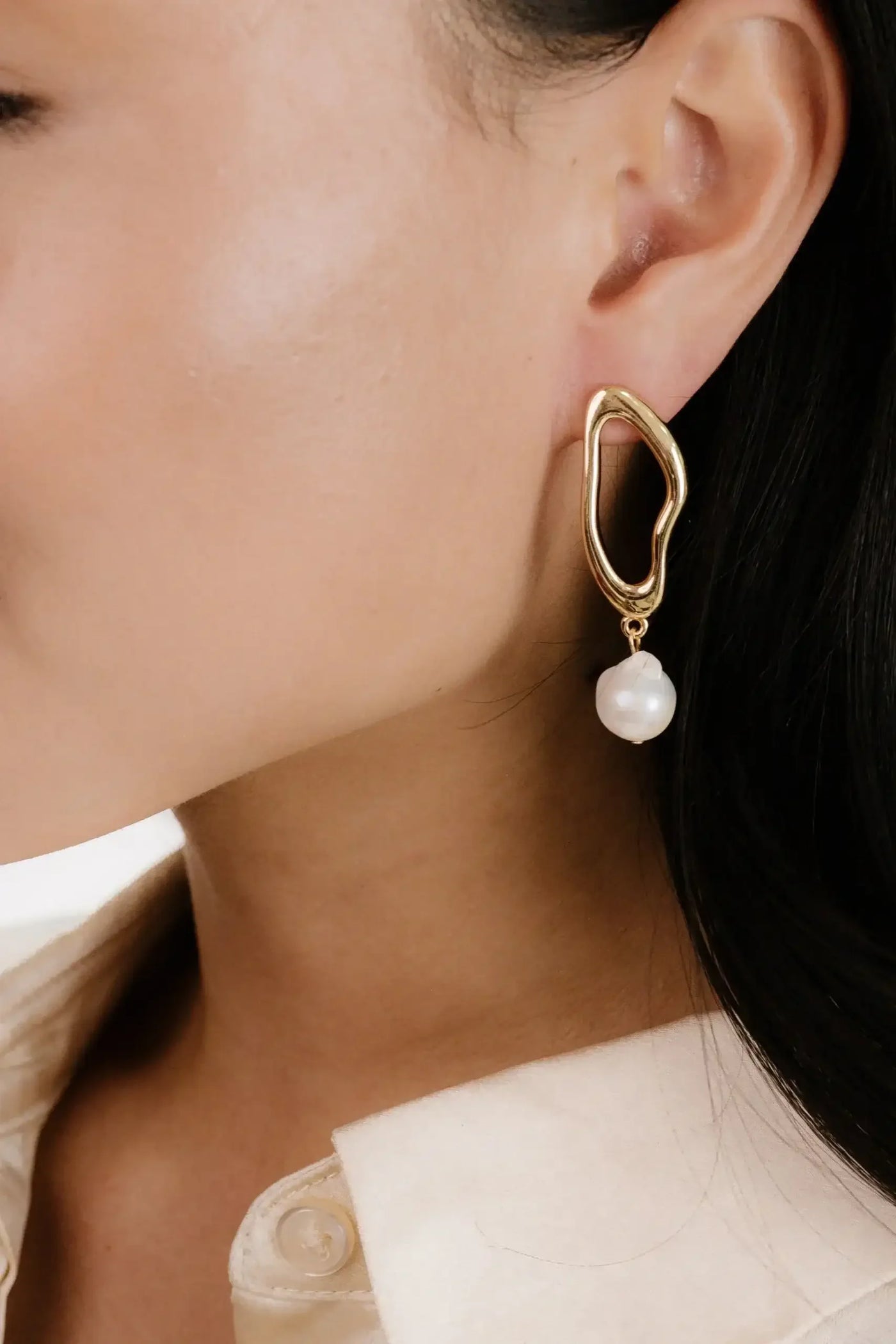 A gold abstract-shaped earring with a hanging pearl is worn on the ear of a woman. She has smooth skin and is dressed in a light-colored, collared shirt.