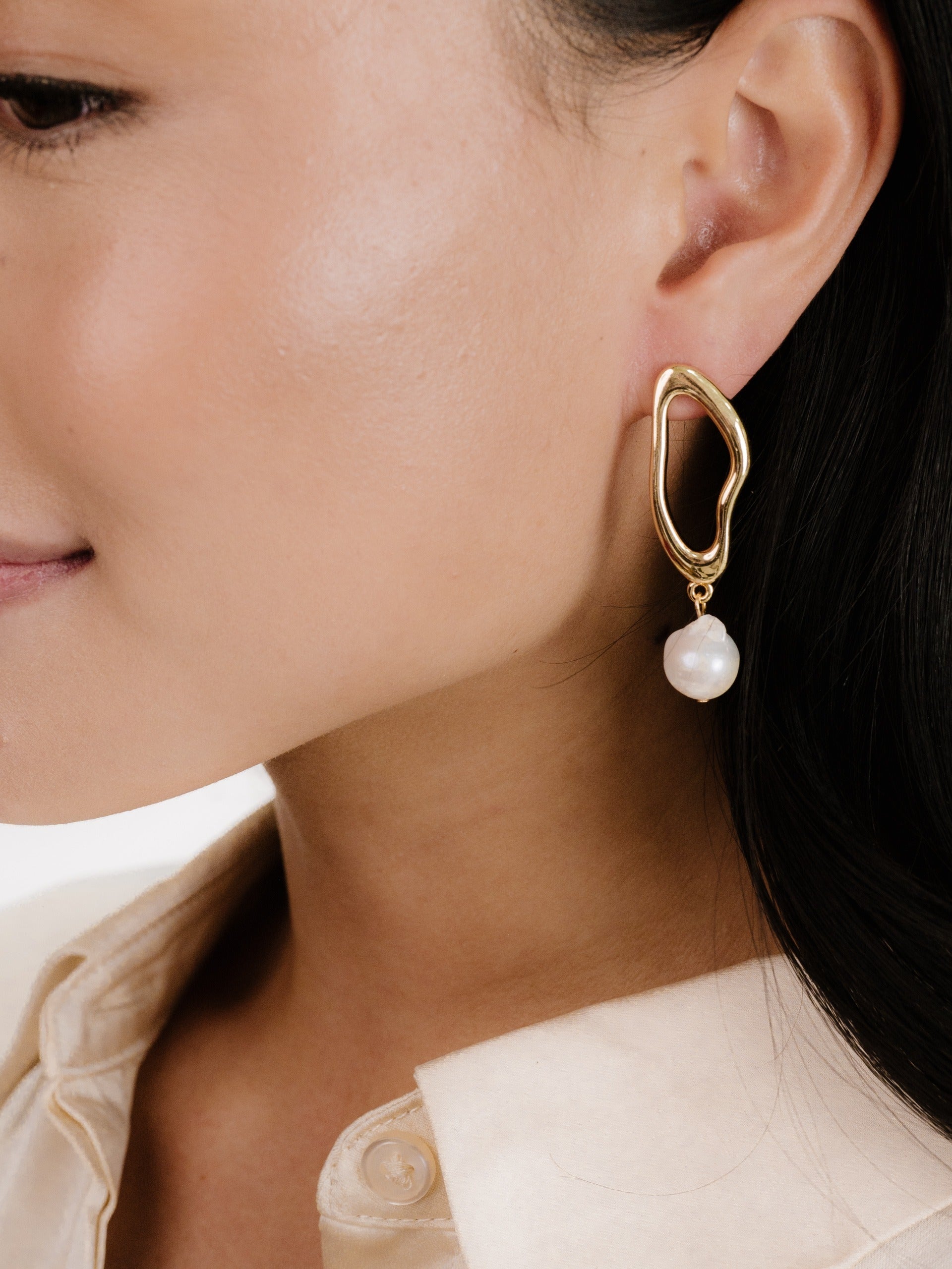A gold abstract-shaped earring with a hanging pearl is worn on the ear of a woman. She has smooth skin and is dressed in a light-colored, collared shirt.