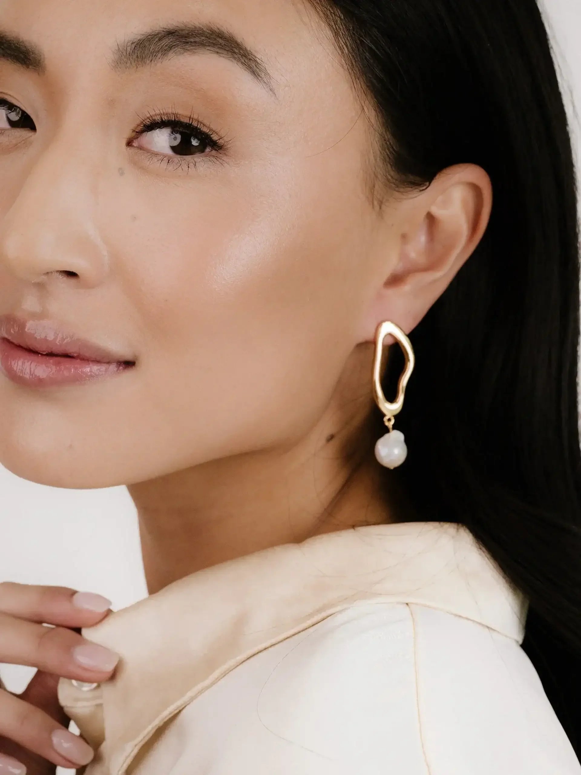 A woman gently touches the collar of her pale garment, showcasing a stylish gold earring with a pearl, set against a neutral backdrop that highlights her clear skin and elegant features.