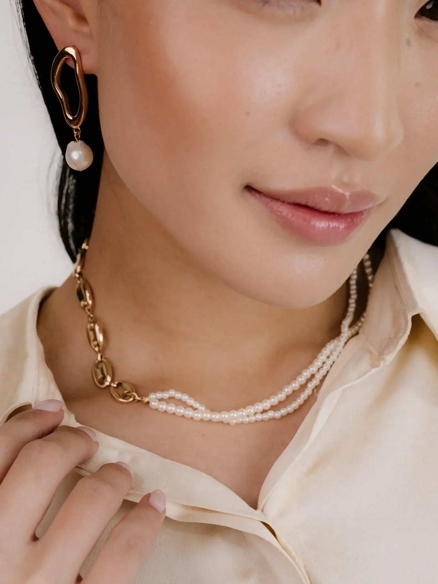 A close-up view of a woman's face showcases her wearing a unique gold earring with a pearl and a layered pearl necklace. She gently touches her collar, highlighting the elegant jewelry against a light-colored silk blouse.