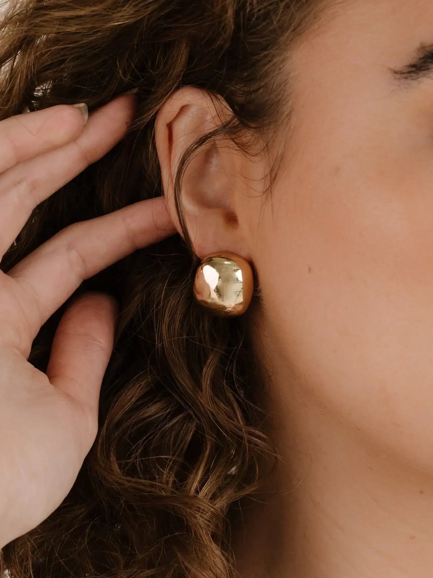 A hand gently touches a woman's ear, highlighting a large, shiny gold earring. The background features softly blurred hair, emphasizing a casual, intimate moment focused on the earring's design.