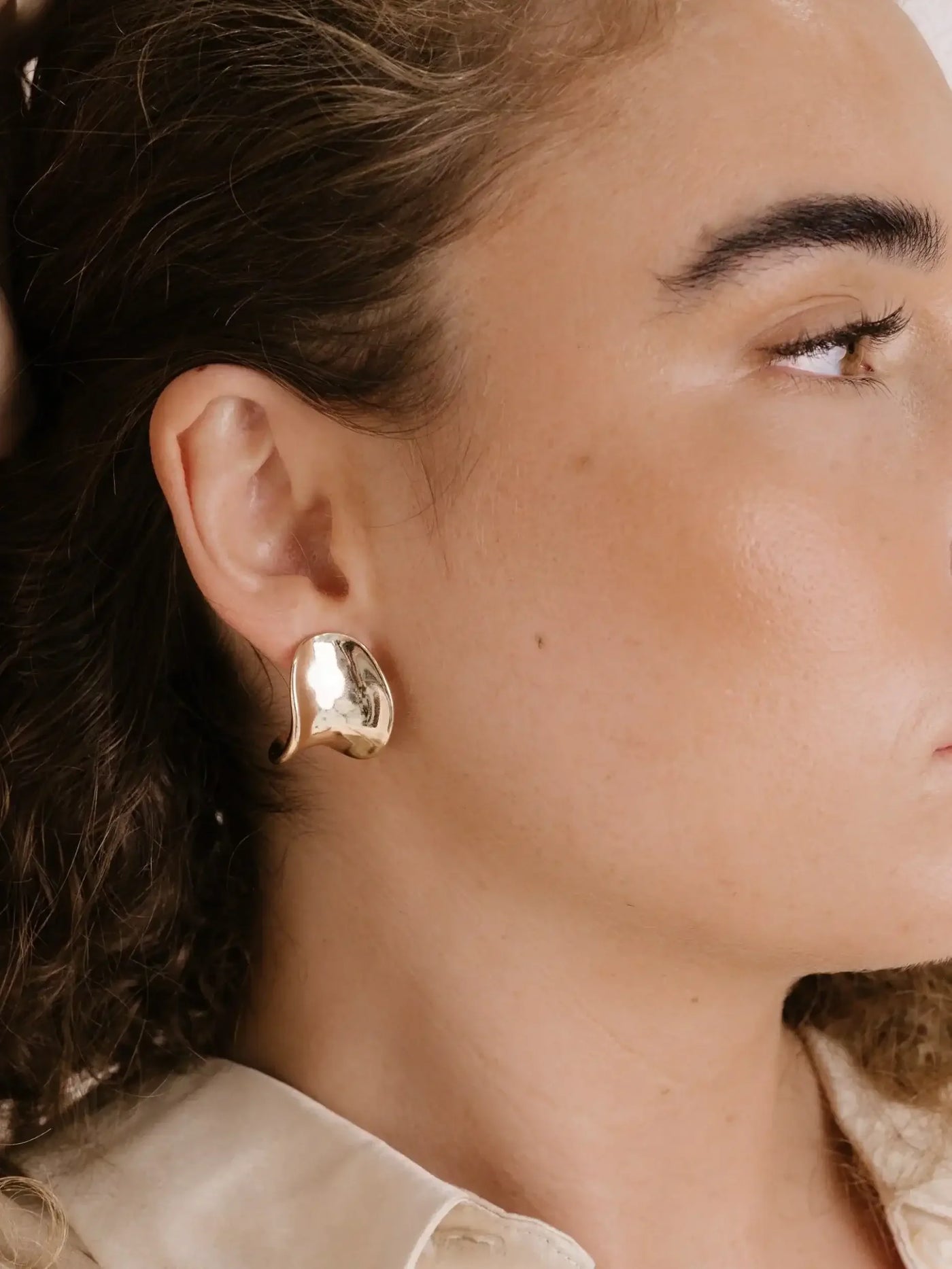 A close-up of a personâ€™s ear adorned with a shiny, abstract gold earring. The individual has curly hair, and they are wearing a light-colored blouse. The skin appears smooth and glowing.