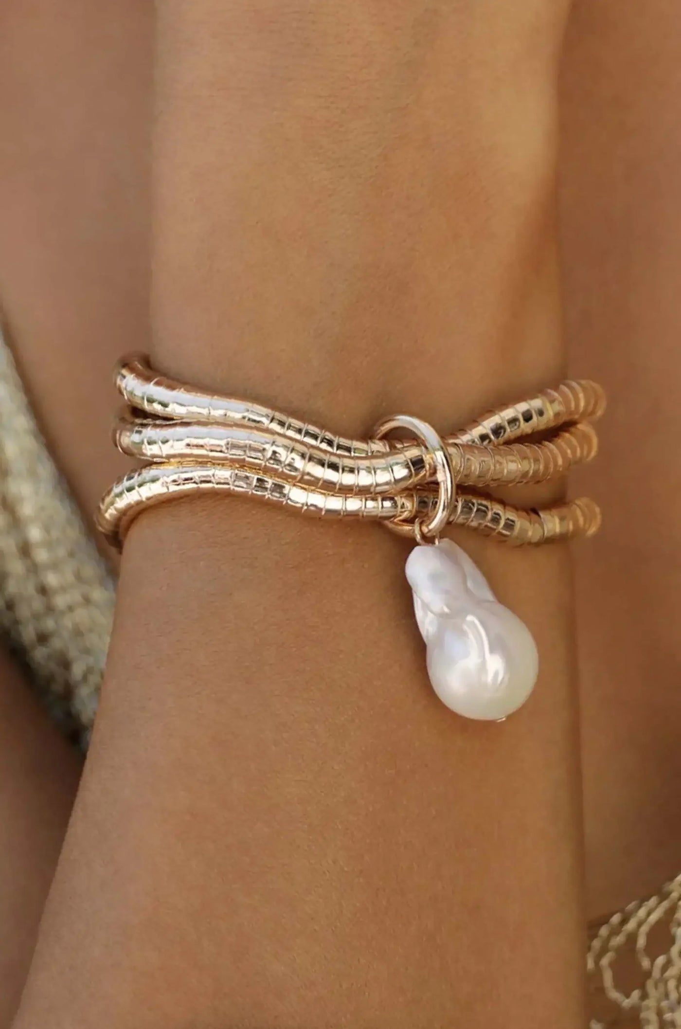 A golden, coiled bracelet with three strands encircles a wrist, featuring a dangling, irregularly shaped pearly charm. The background is blurred, suggesting a warm, casual setting.