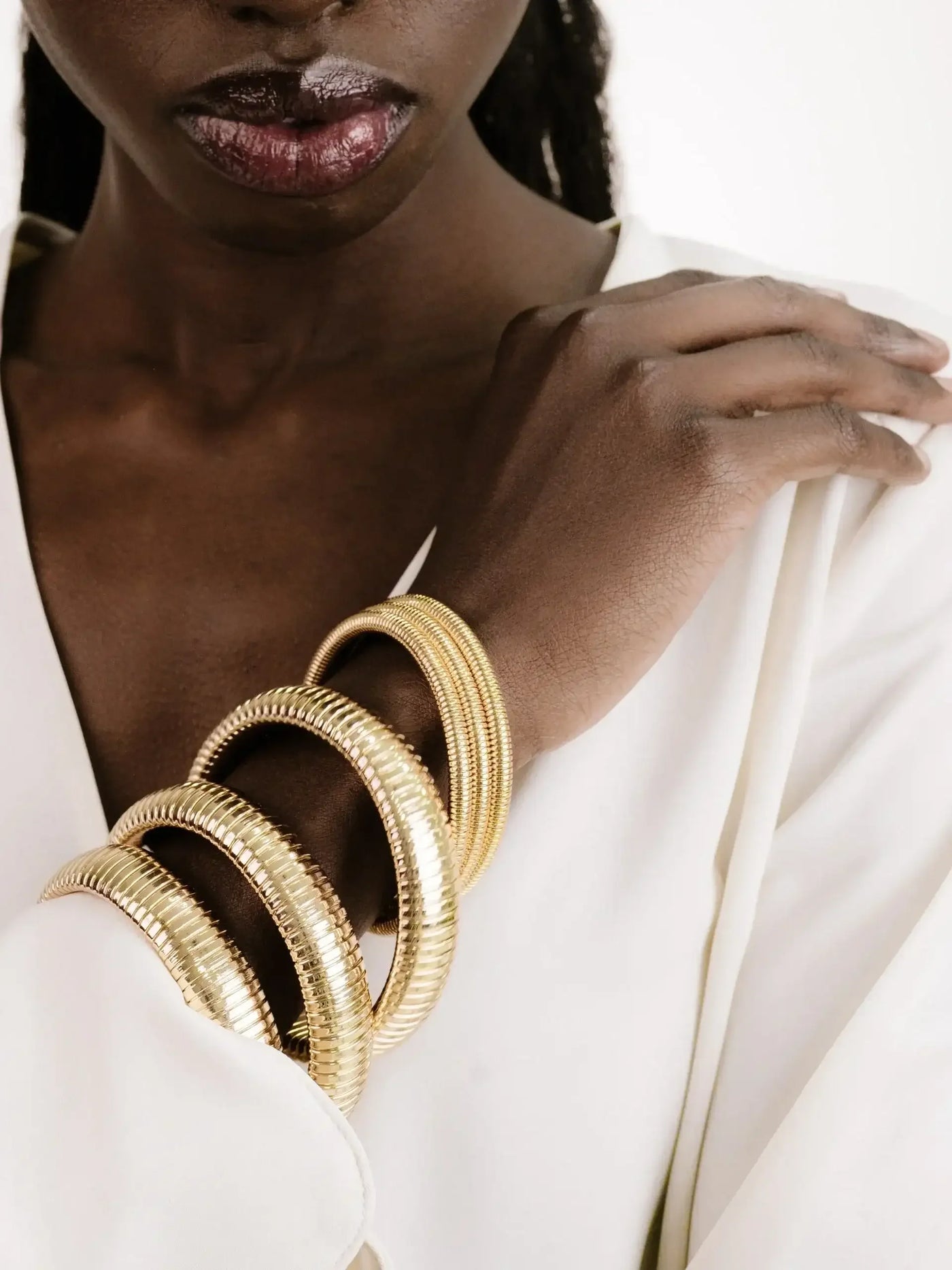 A hand with stacked gold bangles rests against a white garment, showcasing elegant jewelry against a dark skin tone, while lips glisten softly in the background.
