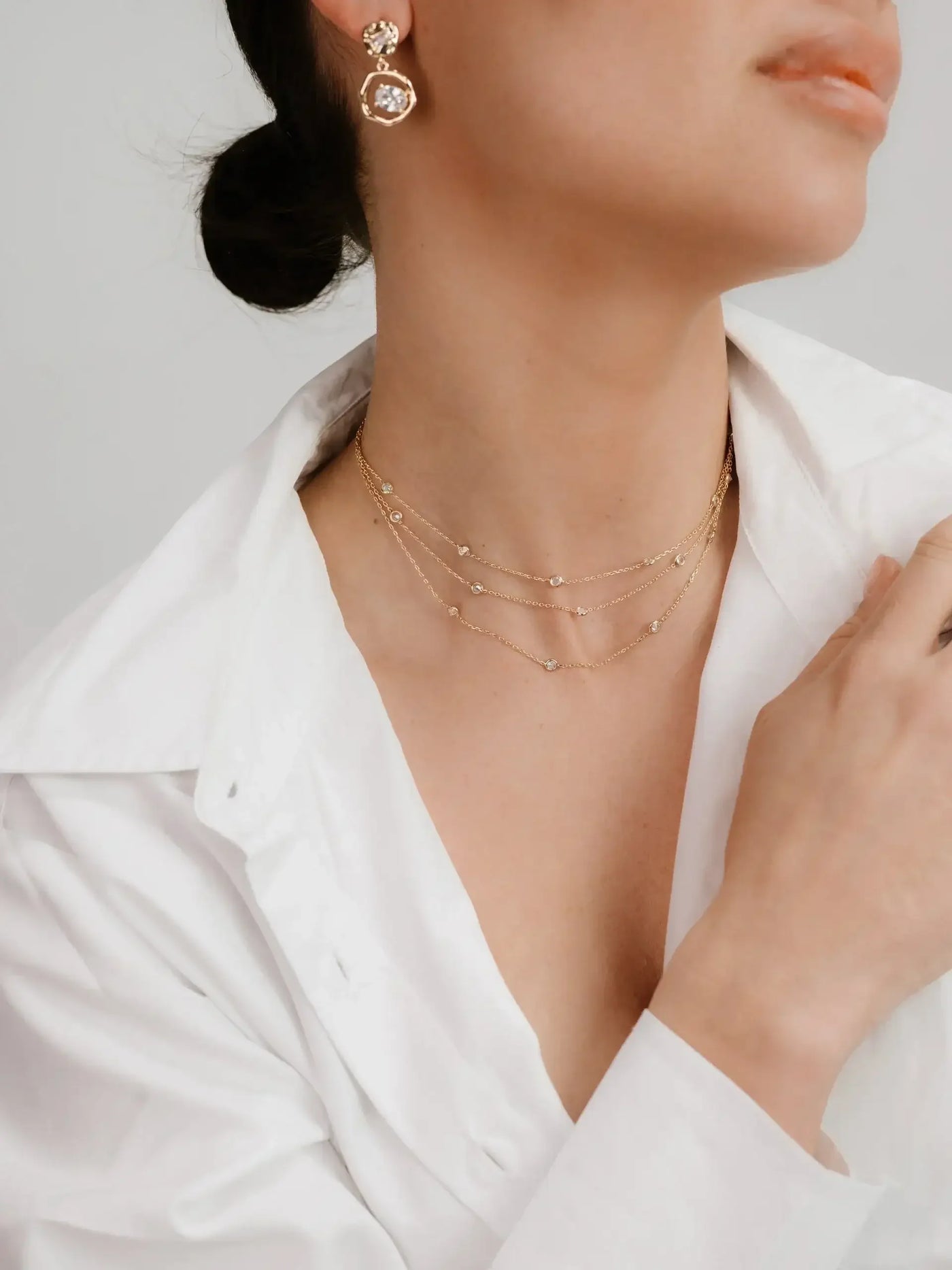 A woman poses with her head slightly turned, showcasing layered gold necklaces and elegant earrings while wearing a white shirt. The background is neutral and unobtrusive.
