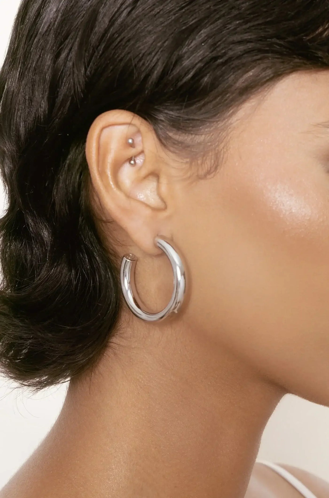 A close-up of an ear adorned with a large, shiny silver hoop earring. The setting features smooth skin tones and soft lighting, highlighting the earring's reflective surface against the textured hair.