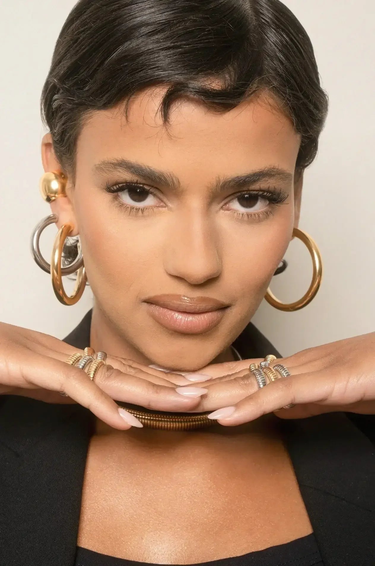 A woman with short, well-groomed hair poses confidently, resting her chin on her hands adorned with rings, wearing large earrings and a stylish jacket, against a neutral background.