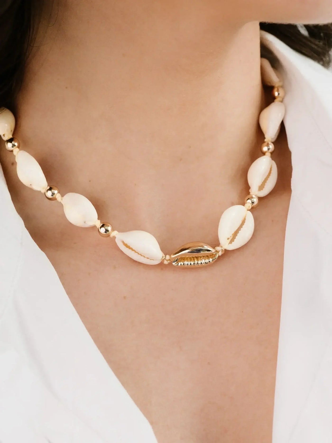 A necklace made of white shells and gold beads adorns a person's neck, resting against smooth skin, with a simple white shirt partially visible in the background.