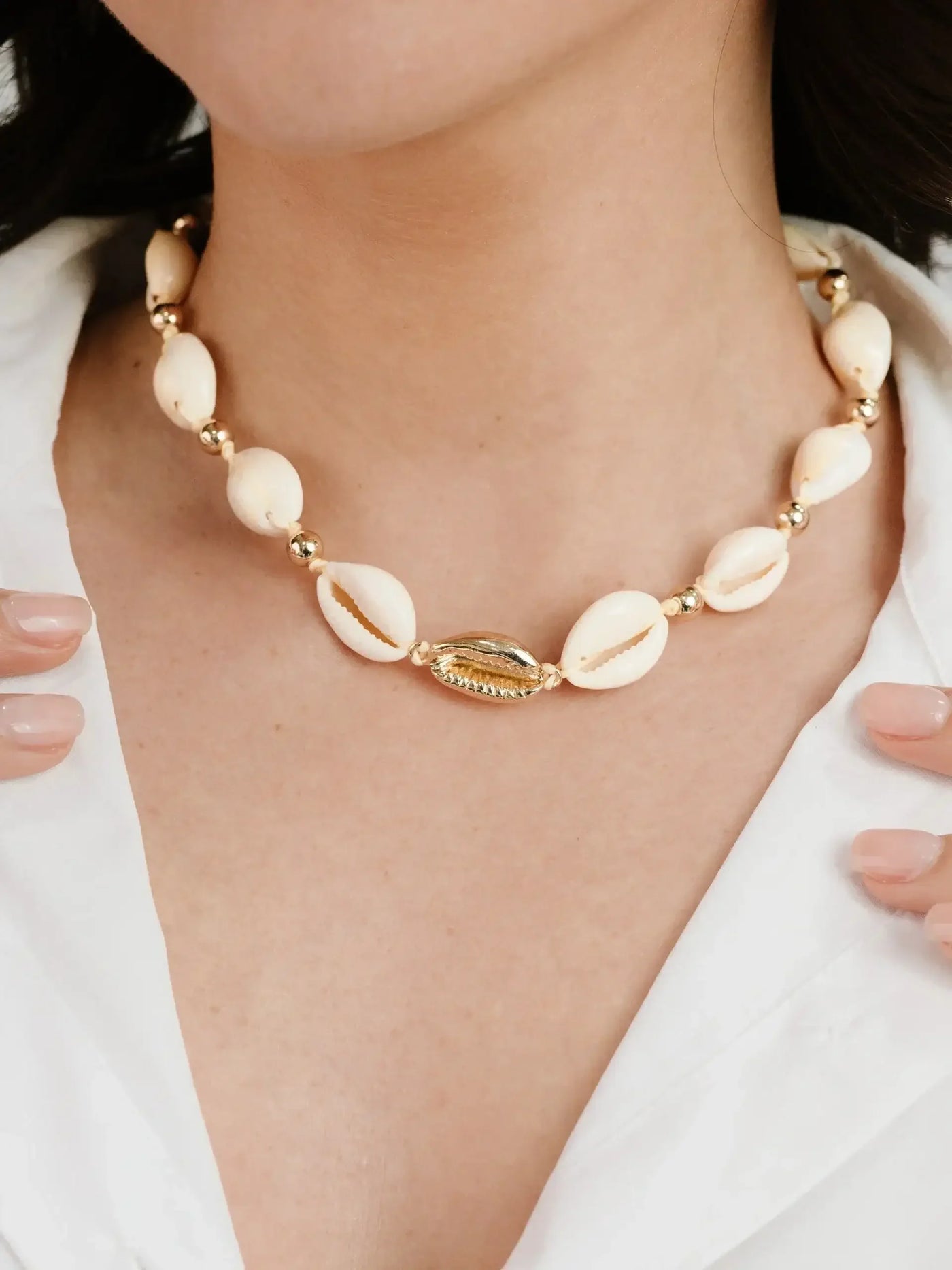A beaded necklace with cream-colored shells and gold accents adorns a person's neck. The individual gently touches the necklace, wearing a white shirt in a light, neutral setting.