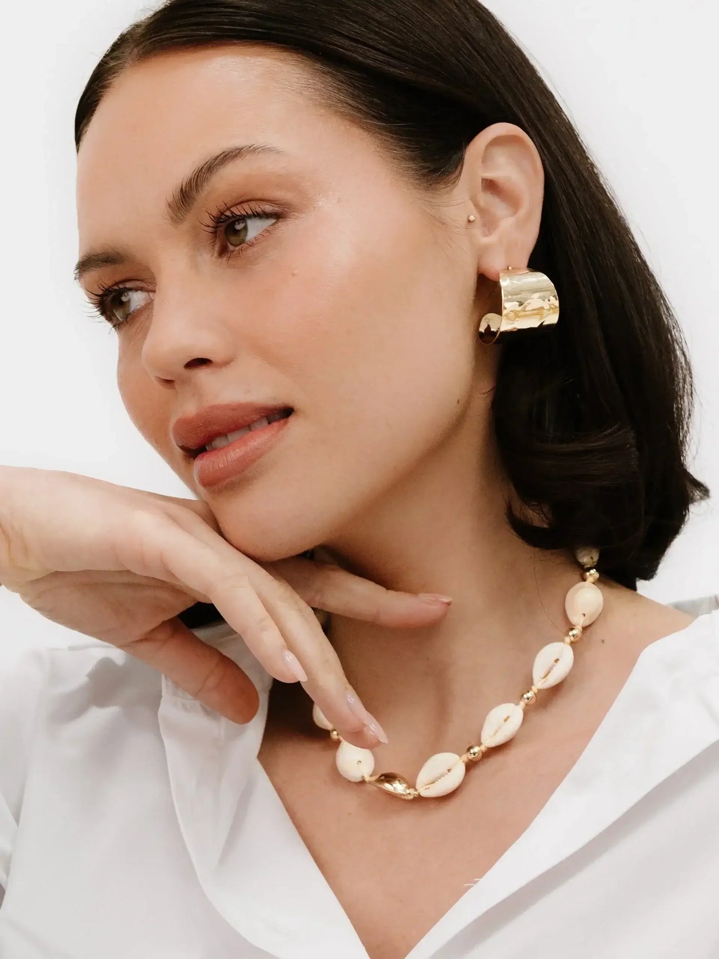 A woman with brown hair and a white shirt poses thoughtfully, resting her hand on her chin. She wears a gold earring and a shell necklace against a plain white background.