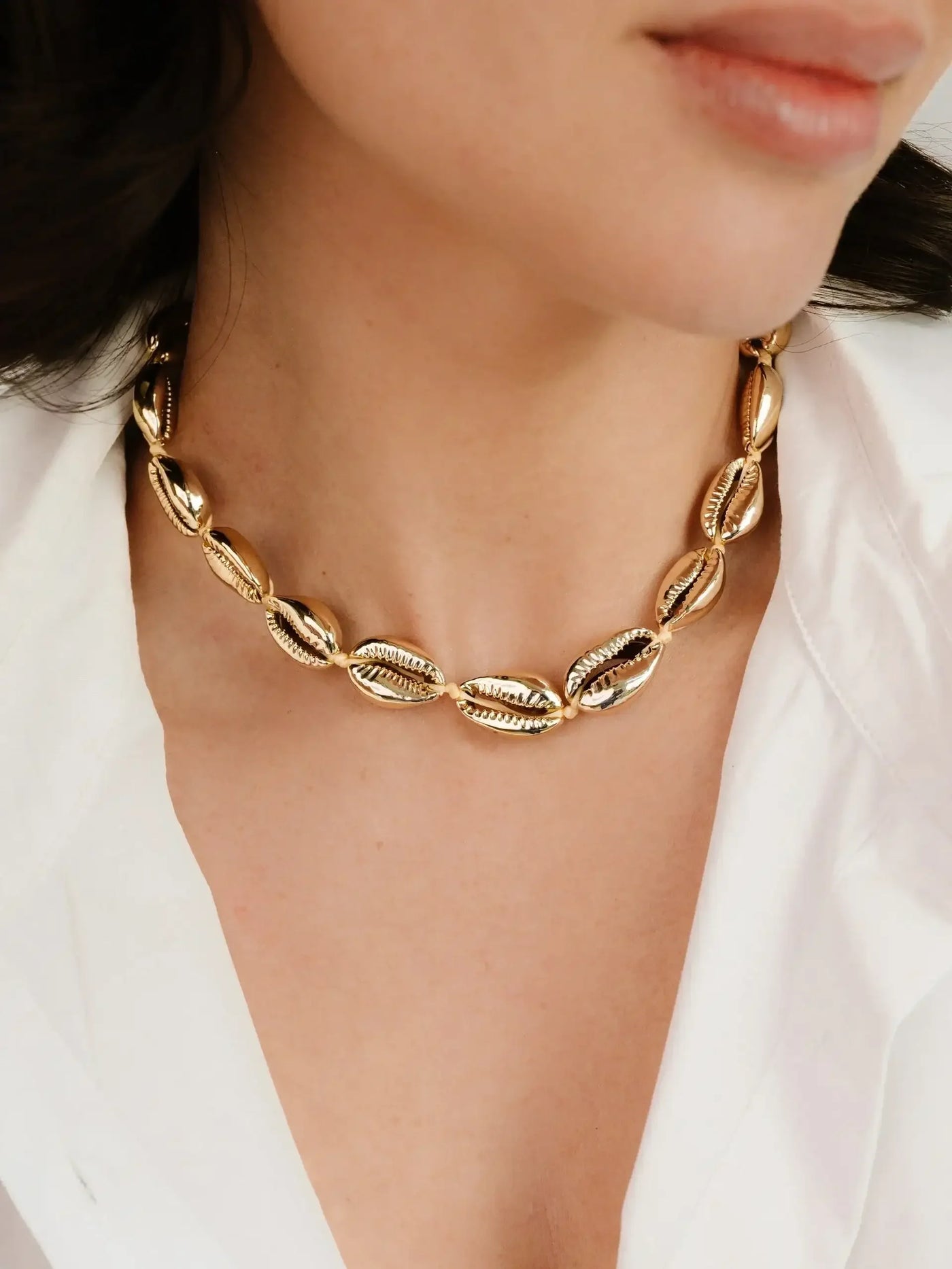 A gold statement necklace, featuring oval links with intricate designs, adorns the neck of a woman in a white blouse. The setting is softly lit, highlighting the jewelry's shine.
