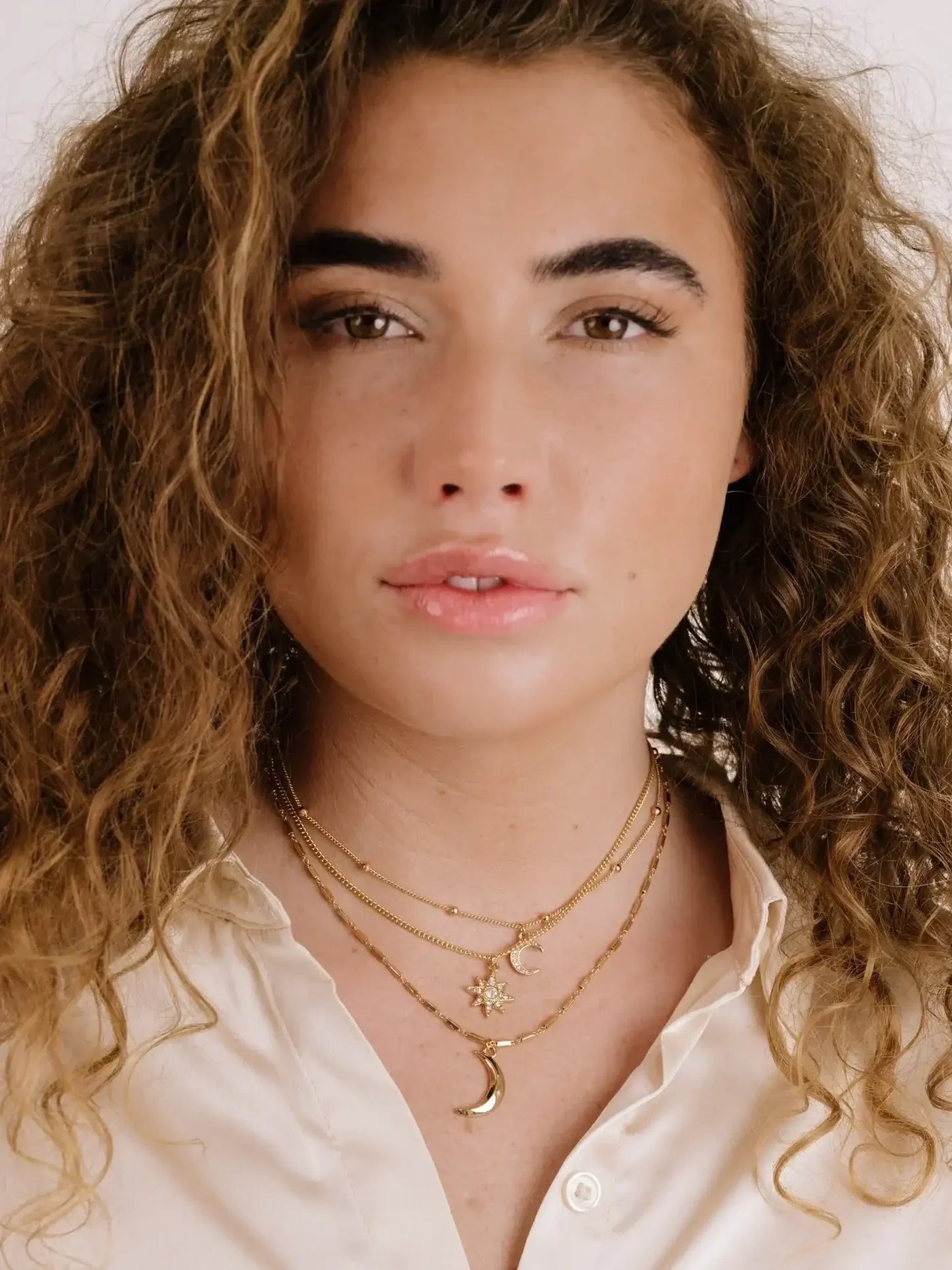 A young woman with curly hair gazes directly at the viewer. She wears a light-colored blouse and layered gold necklaces featuring moon and star charms, set against a plain background.